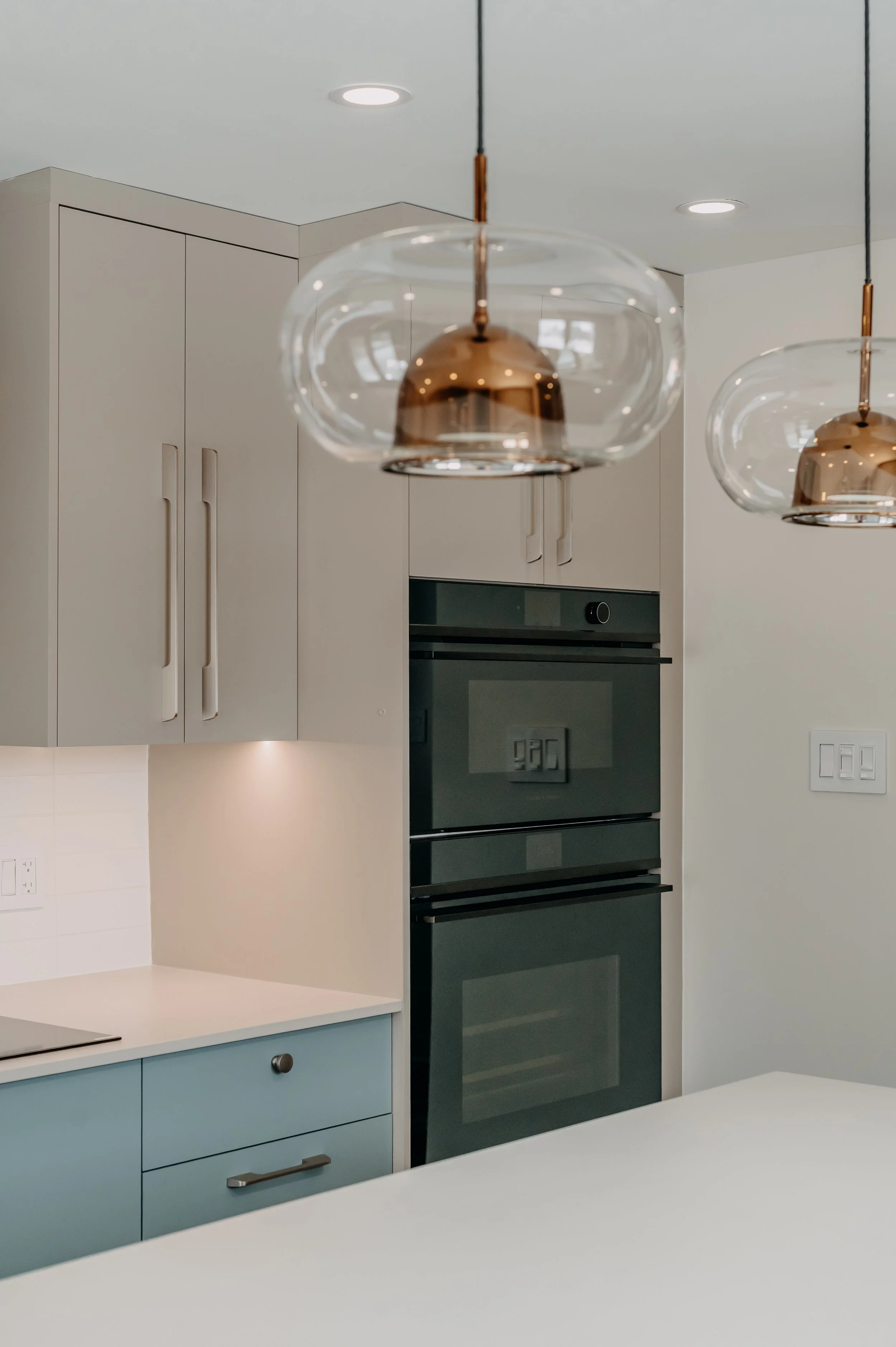 A sleek built-in double oven console sits beneath brass pendant lighting, an ABtelier Design detail blending sunny character with everyday function in this blue kitchen.