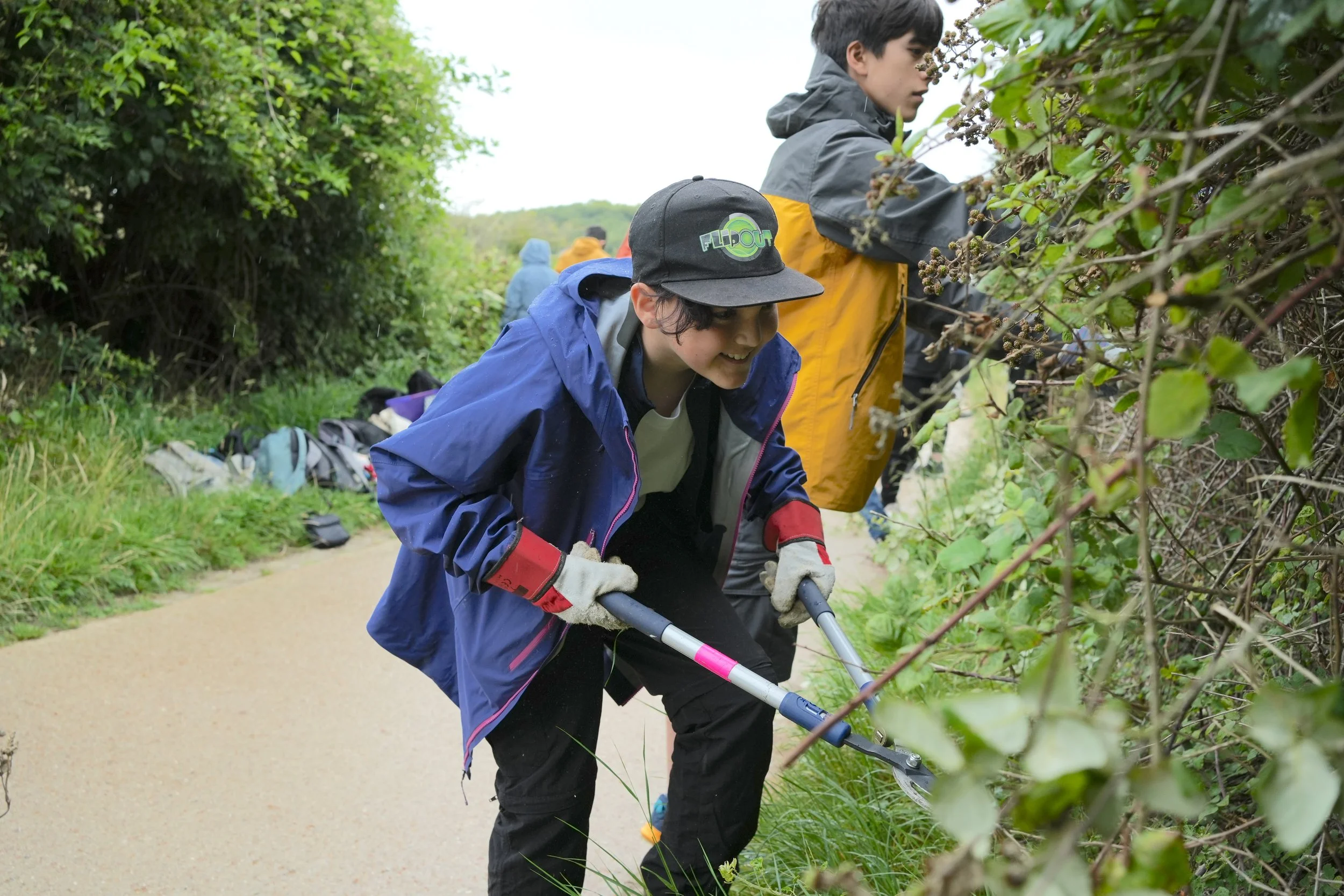 Connor leans forward holding loppers, he is trimming back bramble as part of a hands-on conservation activity with green foliage behind