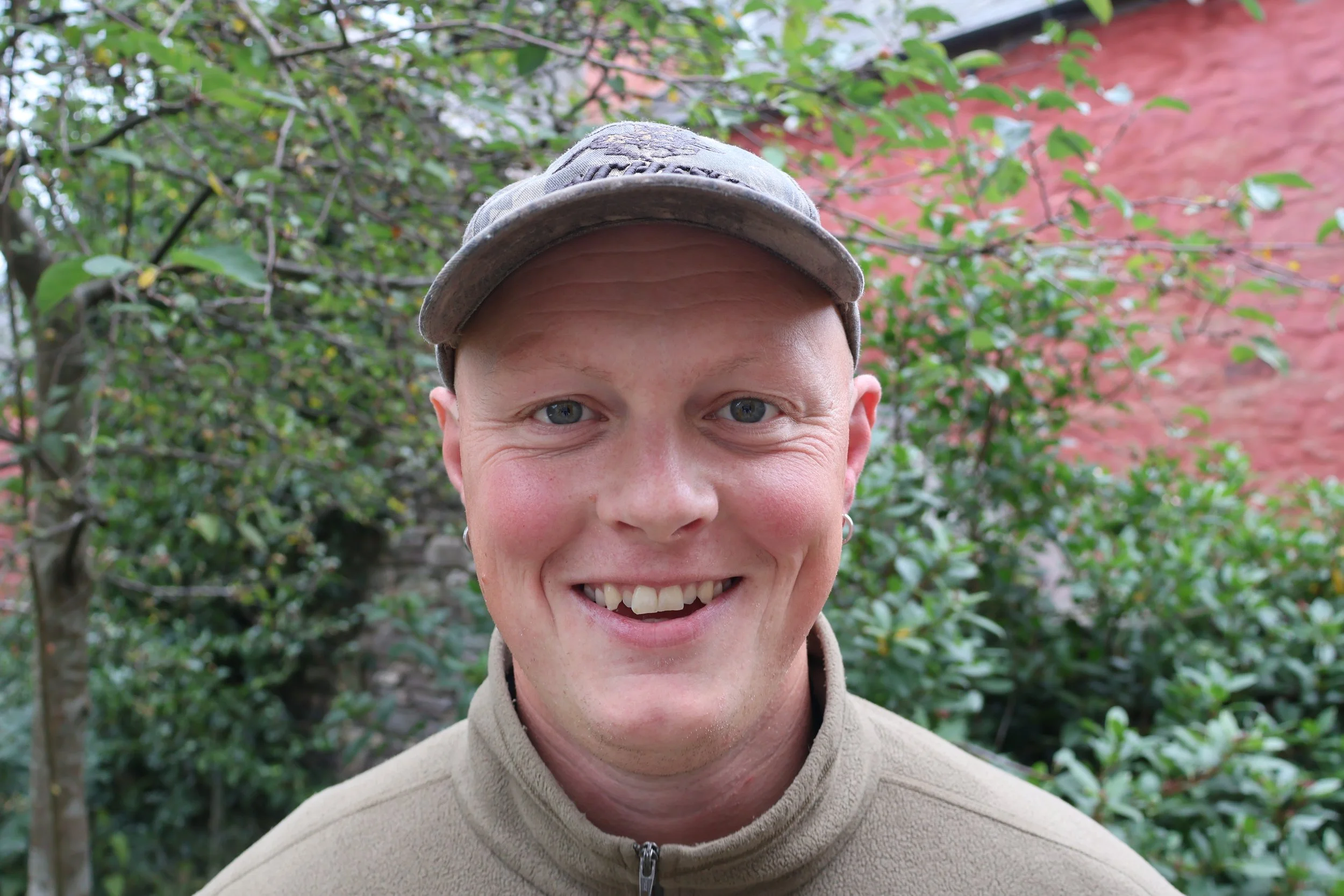 A smiling man wearing a gray beanie and a black waterproof jacket, standing outdoors next to a large tree with green leaves, on a grassy hillside with a partly cloudy sky in the background.