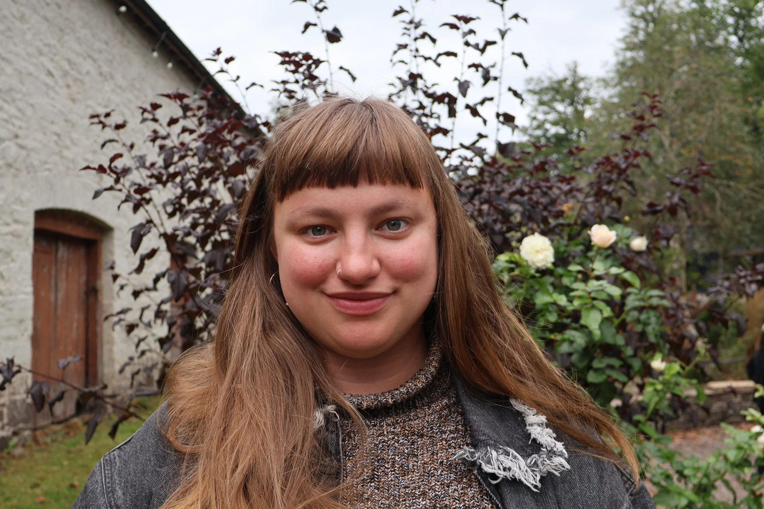A young woman with short brown hair and a nose piercing standing in a grassy field during sunset, wearing a plaid shirt over a graphic t-shirt and denim overalls.