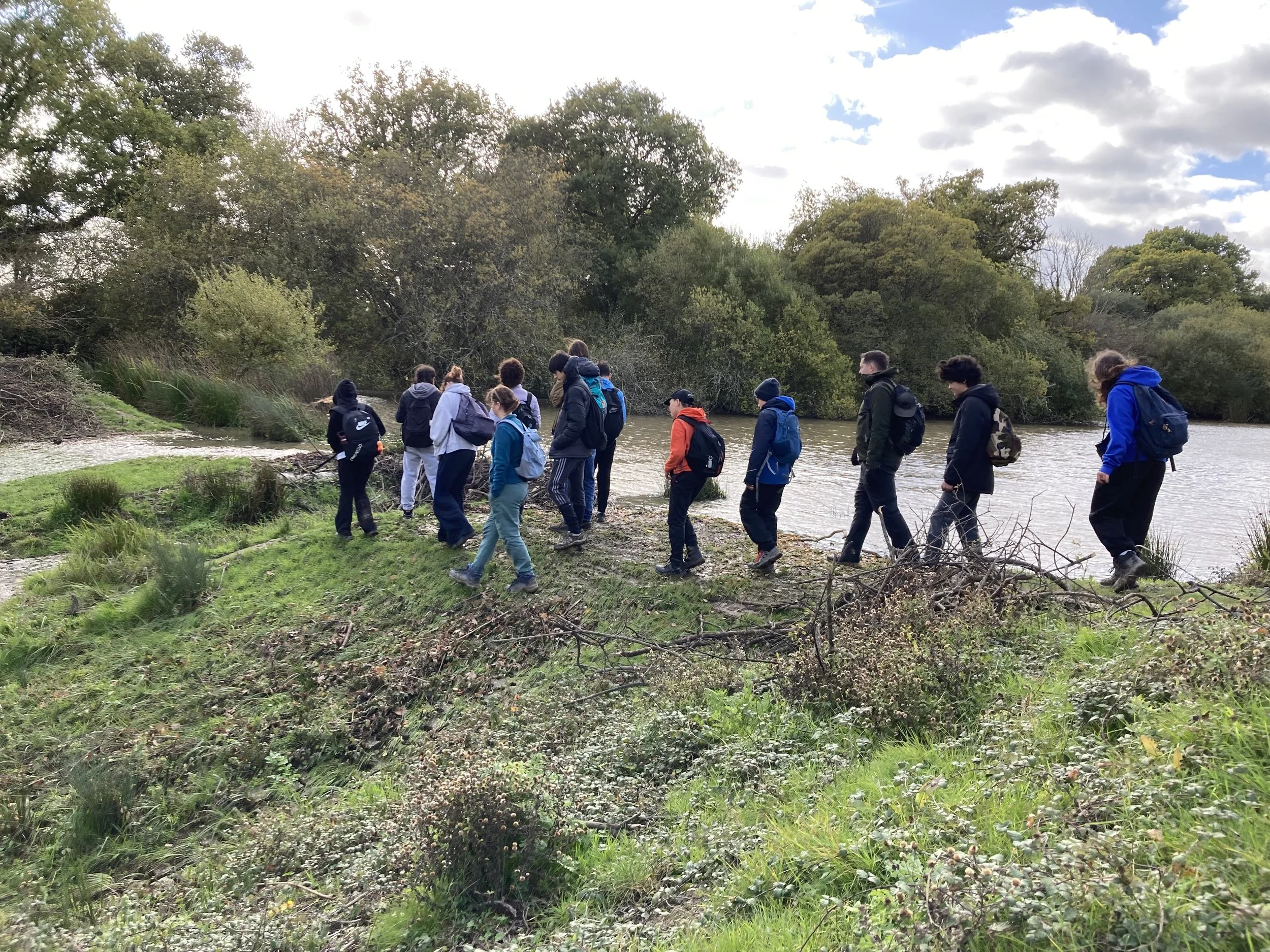 A group of young people walk in nature with trees and a pond behind them.