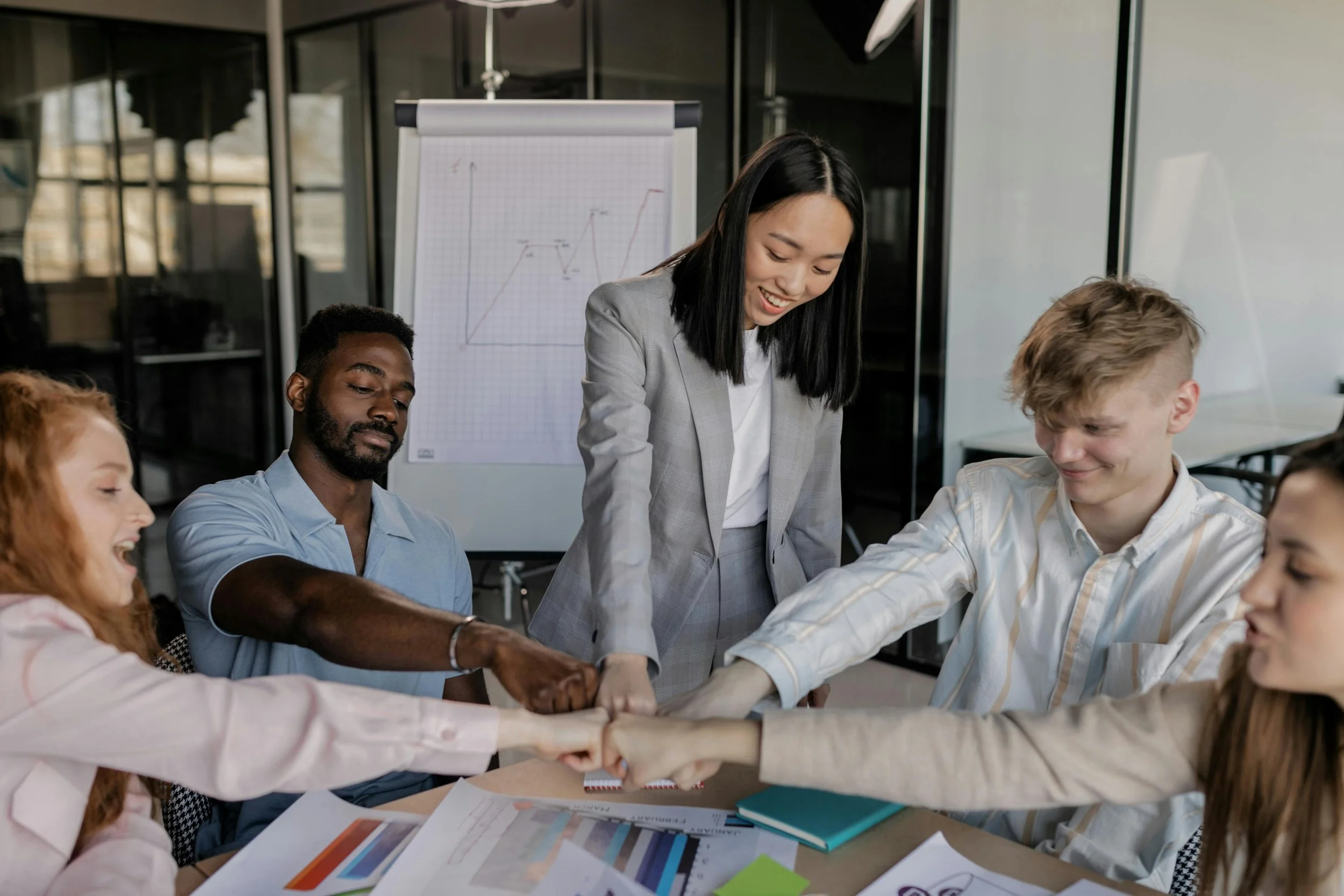 A diverse group of five people in an office room, joining their hands in a team gesture of unity, with a woman in a gray blazer smiling in the center, and a chart with a rising graph in the background.