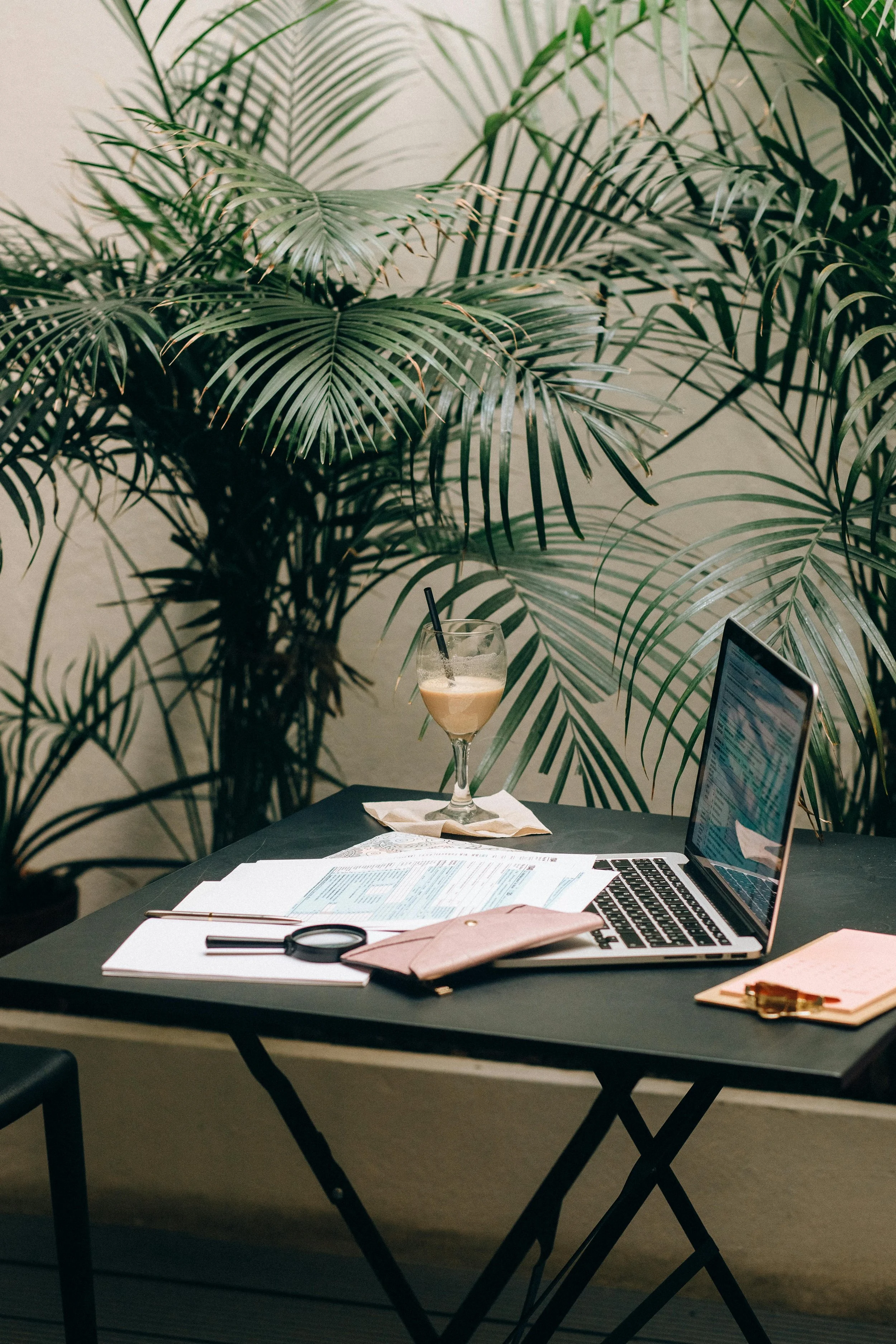 A workspace with a laptop, papers, a magnifying glass, a pink wallet, and a clipboard on a black table, with large green tropical plants in the background, and a glass of iced coffee or tea with a straw.