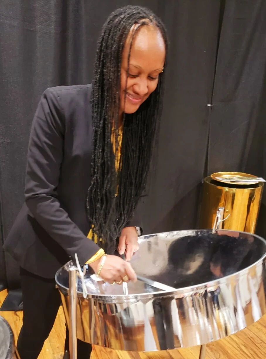 Woman playing a polished steel pan drum onstage against a black curtain.