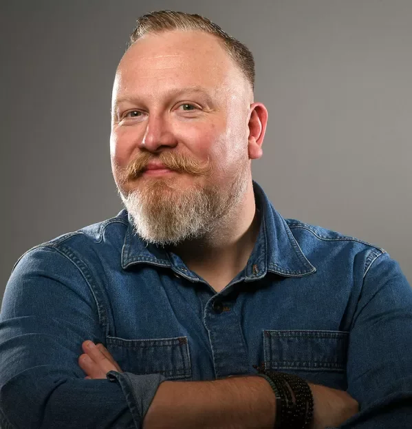 Gareth Conyard, A man with a beard and short hair smiling confidently, wearing a denim shirt, standing against a plain grey background.