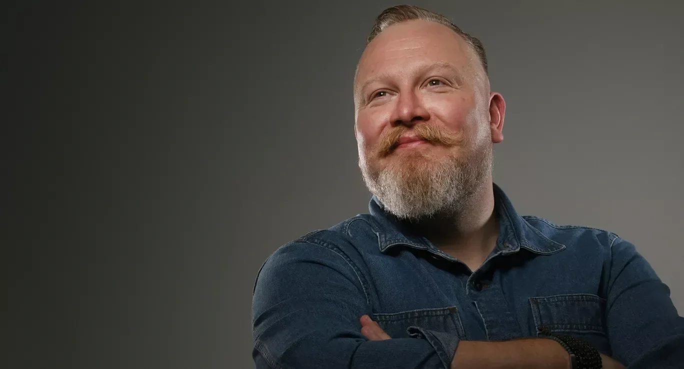 Gareth Conyard, a with a beard and moustache, smiling, wearing a denim shirt and standing against a grey background.