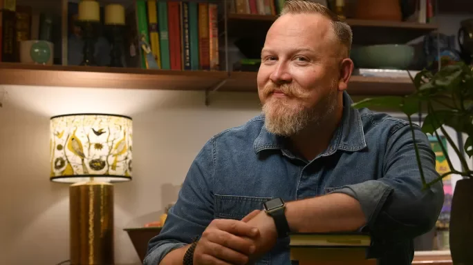 Gareth Conyard, a man with a beard and moustache, wearing a denim shirt, sitting at a desk with a bookshelf in the background.