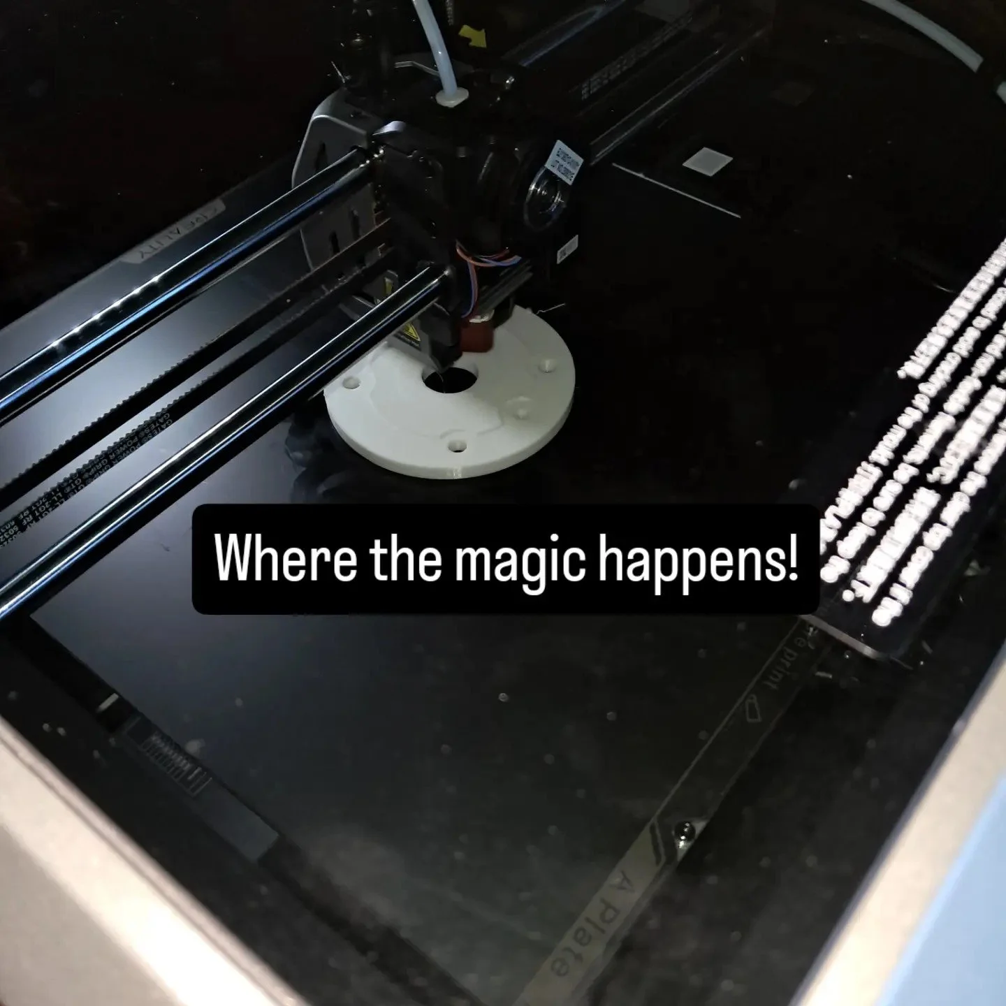 Inside a 3D printer, showing the printing bed, extruder, and filament. The text overlay says, 'Where the magic happens!'.