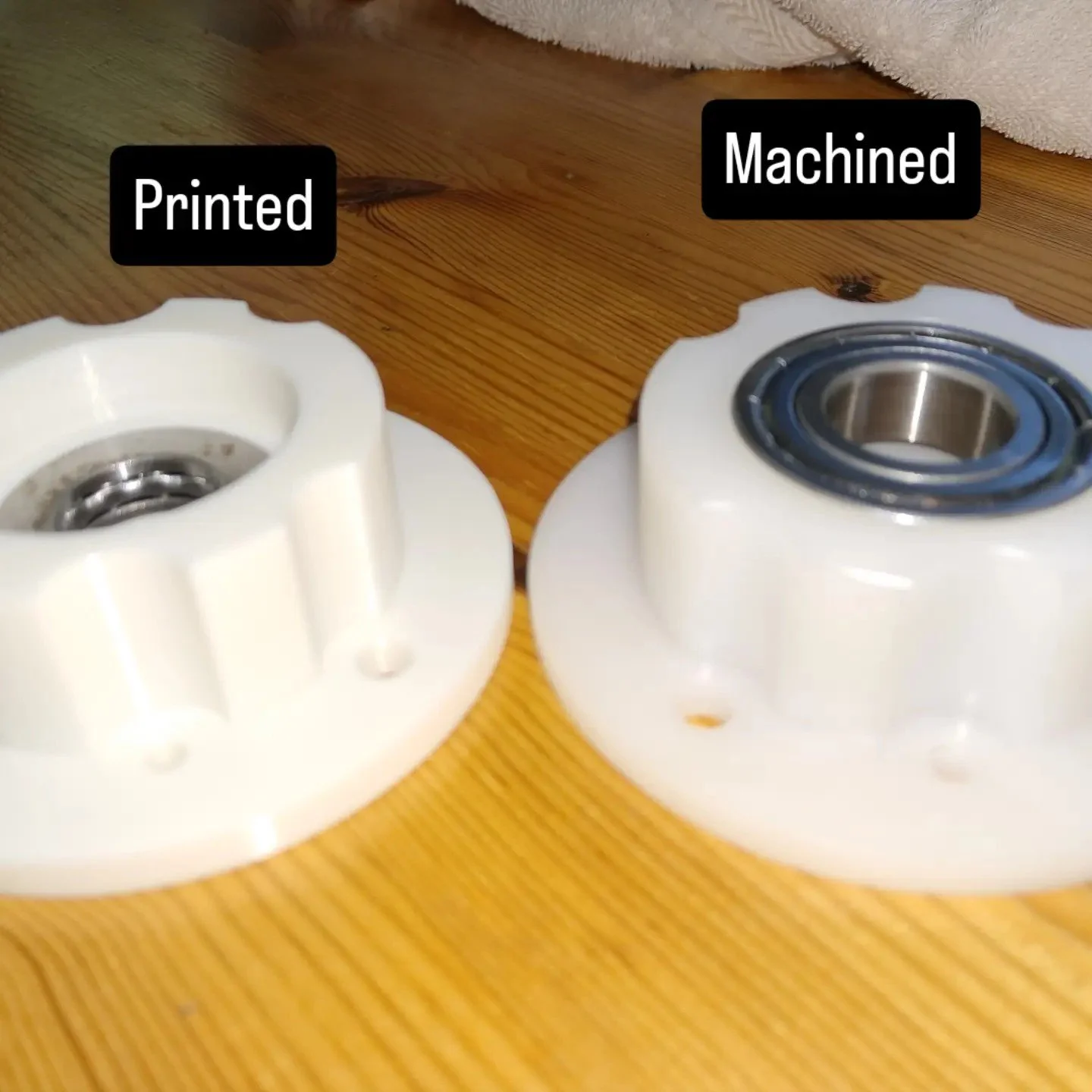 Comparison of two skateboard wheels. The left wheel is labeled 'Printed' and the right wheel is labeled 'Machined.' The wheels are white with metal bearings inside, placed on a wooden surface.