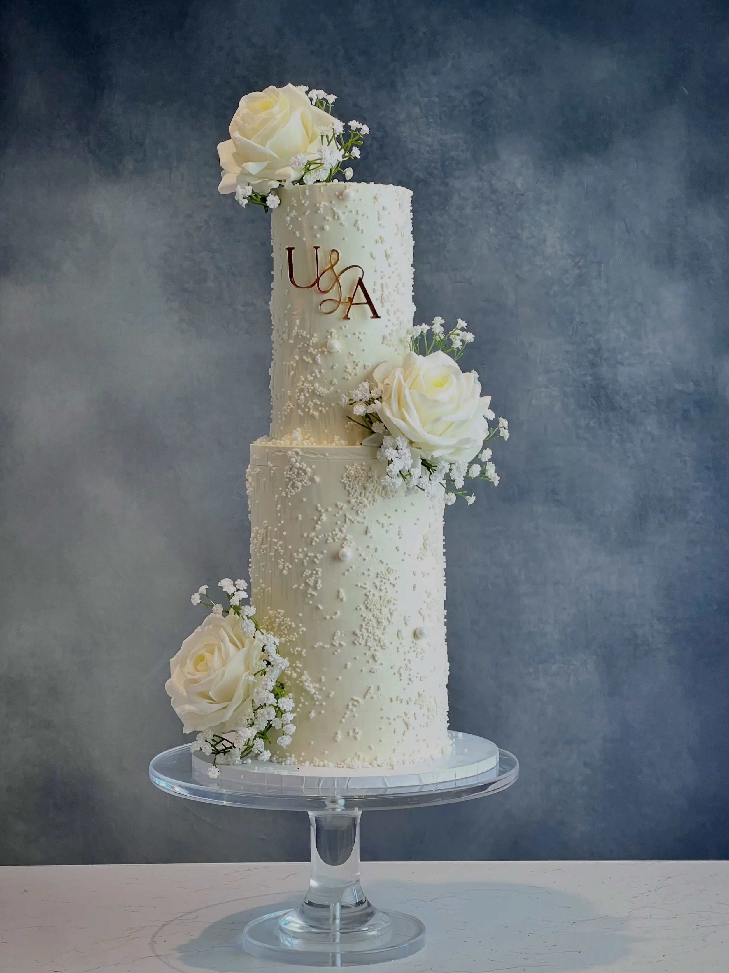 This elegant wedding cake was created for U &amp; A, featuring delicate white roses, intricate pearl detailing and their beautiful personalised initials.

A timeless design, thoughtfully created to complement their special day. 🤍

Planning your big 