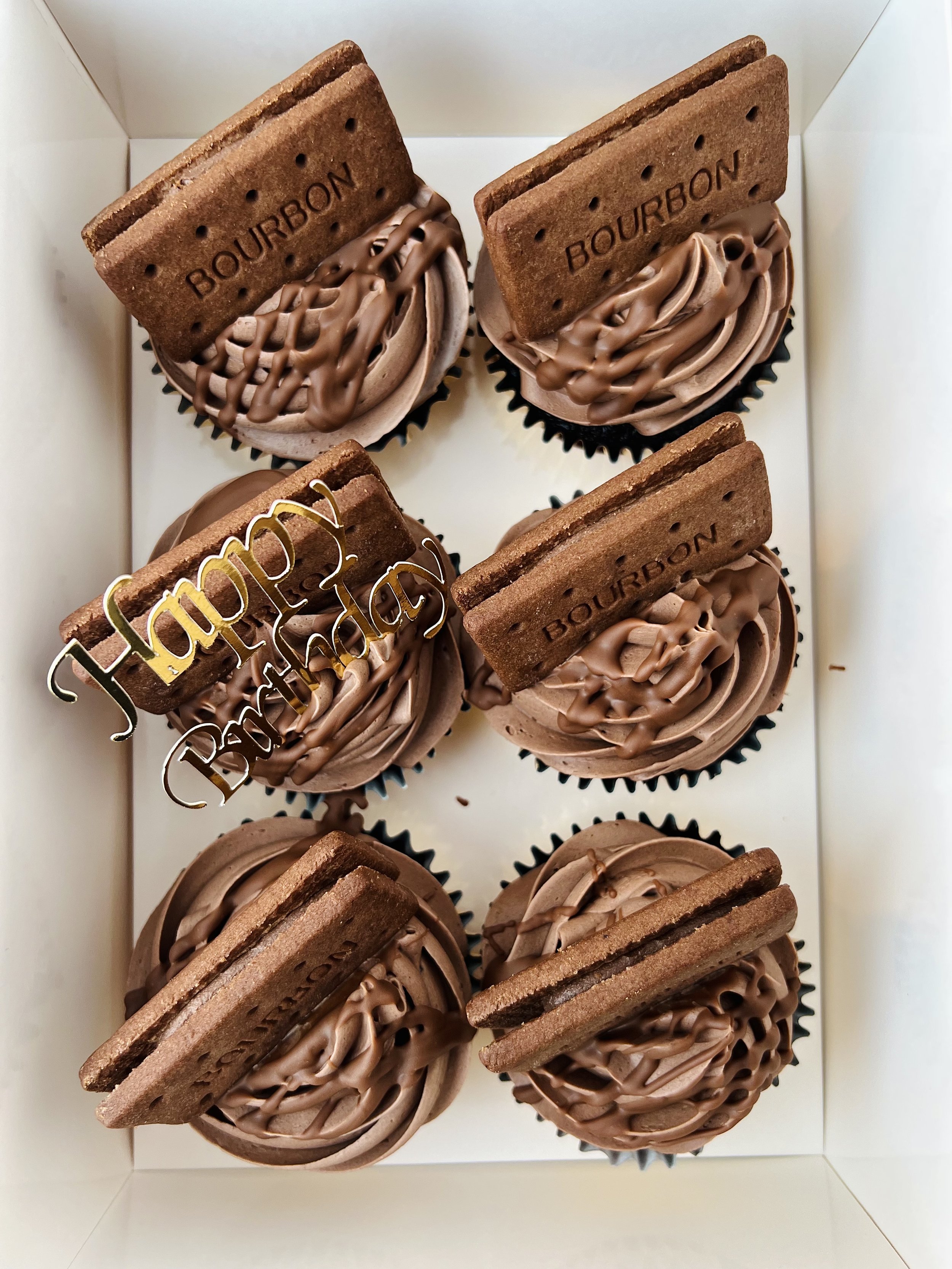 Top view of a Box of 6 Bourbon Biscuit Cupcakes by Sensational Cakes and Desserts with a Happy Birthday Name Charm on one of the biscuits.