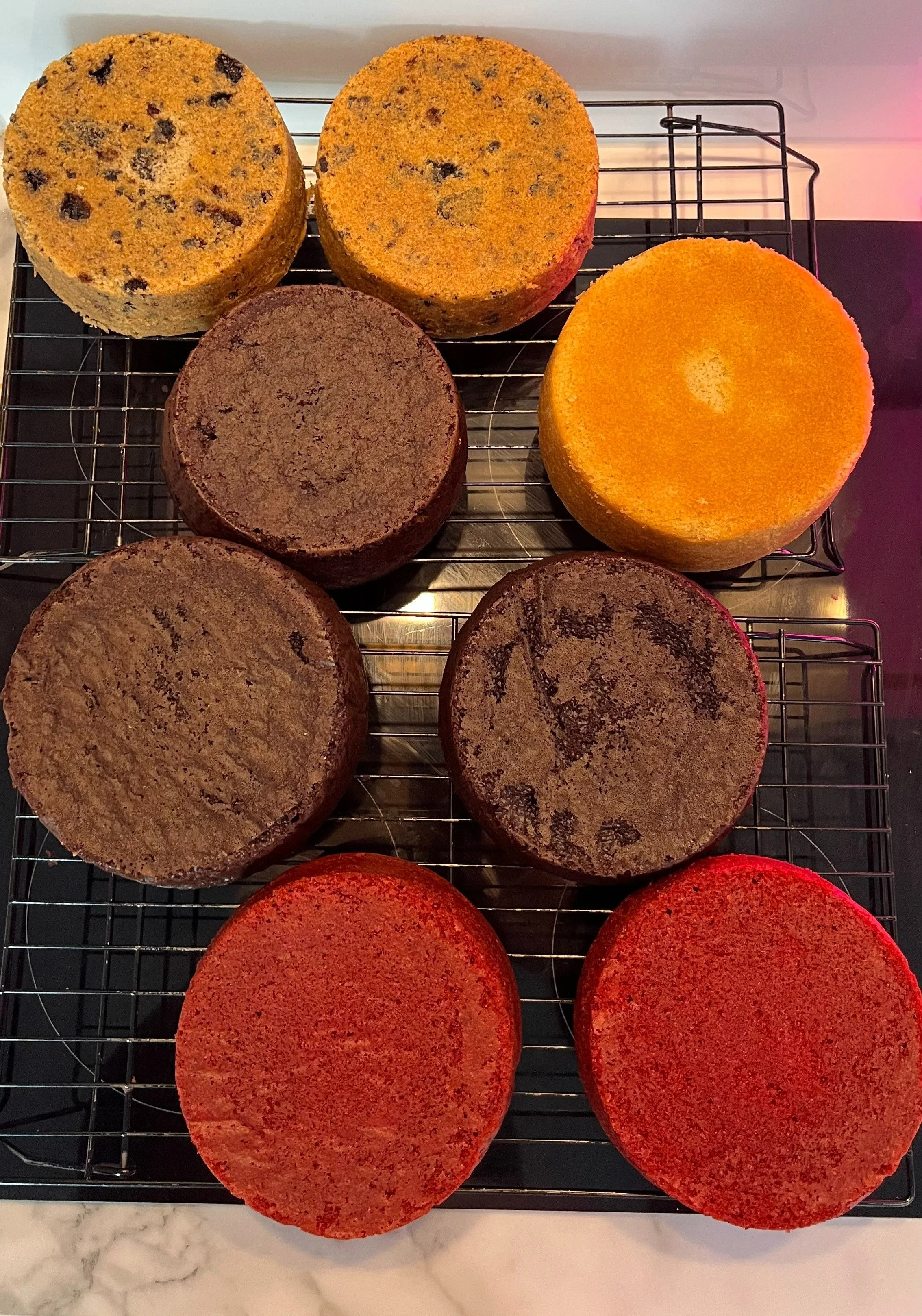 Assorted colorful cake layers cooling on a wire rack by Sensational Cake and Desserts.