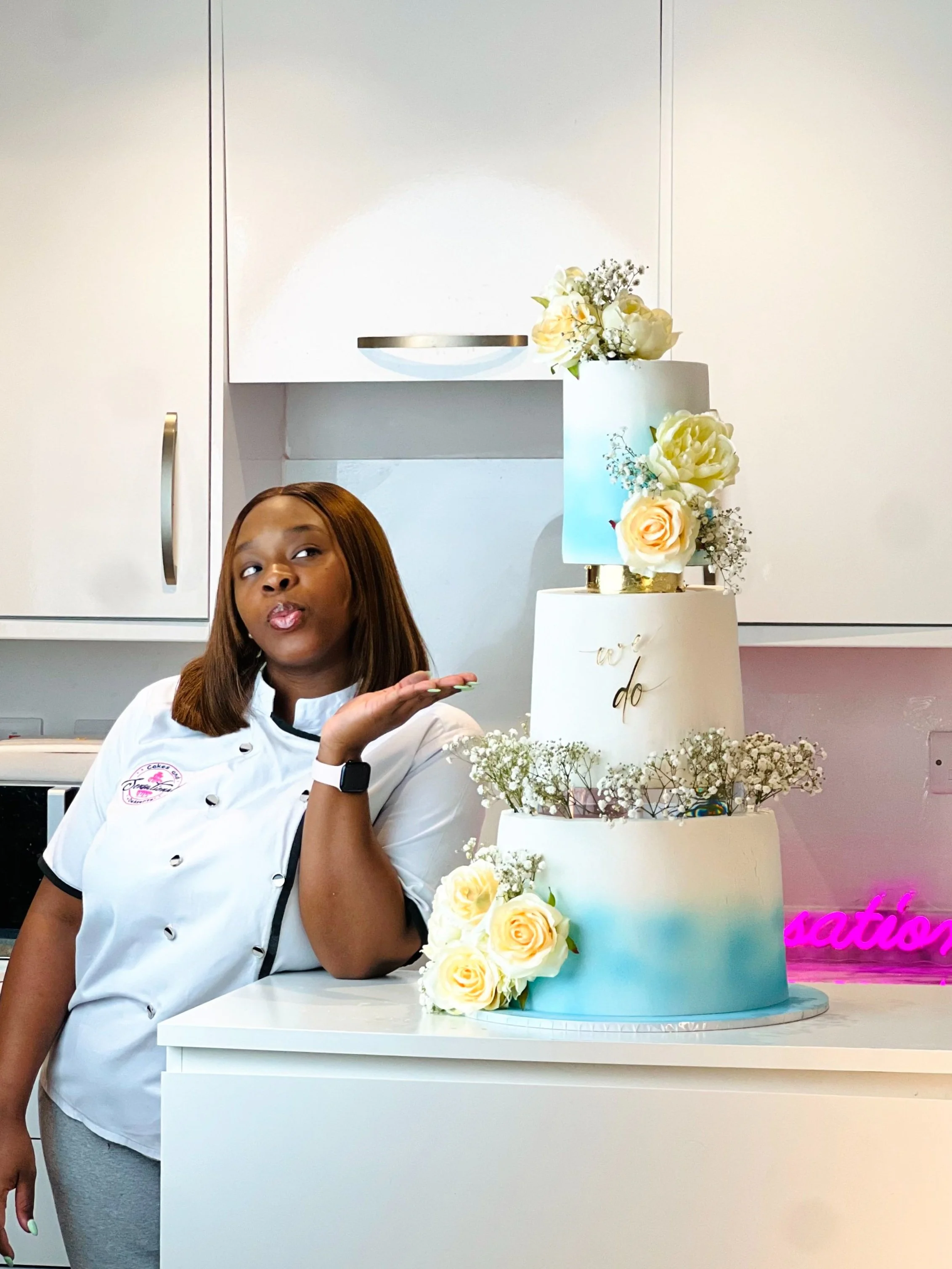 Sope of Sensational Cakes and Desserts in a chef's uniform standing next to a three-tier wedding cake decorated with white roses, baby's breath, and a cake topper with the words 'we do' in gold lettering, in a modern kitchen.