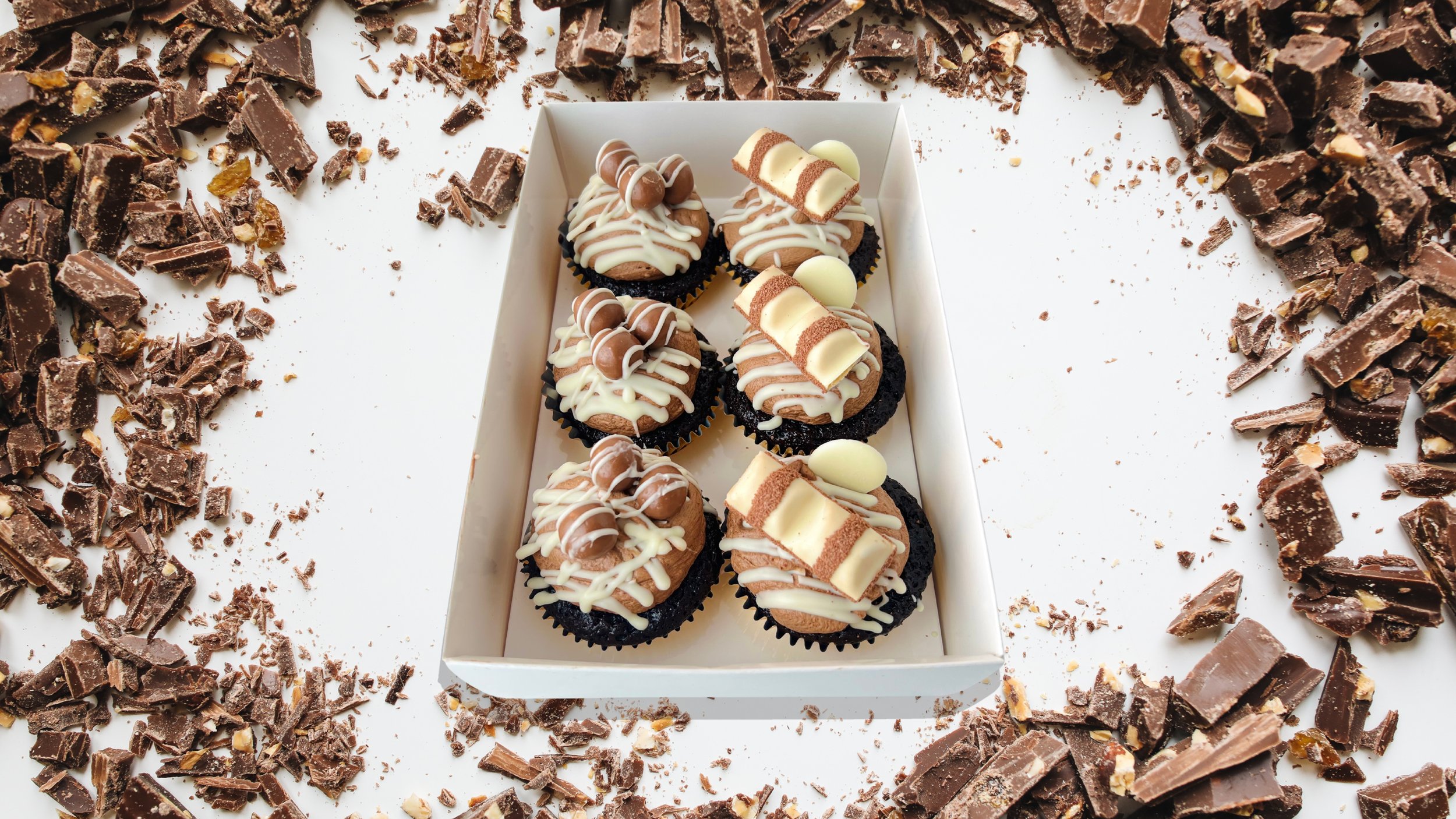 Box 6 Chocolate Cupcakes for by Sensational Cakes and Desserts, topped with Buttercream and Maltersers on the left and Kinder Bueno on the right.