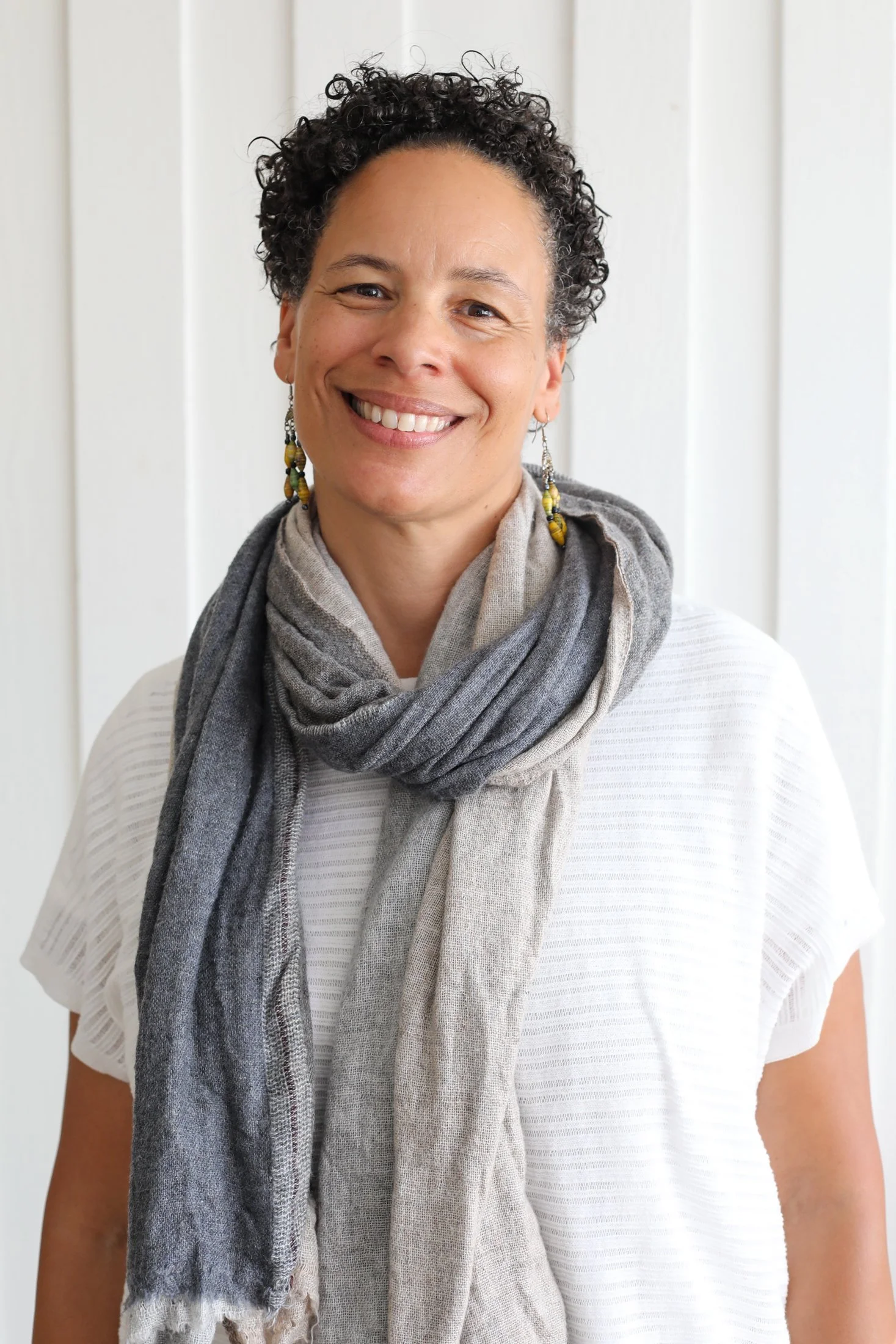Portrait of Victoria, a woman with short curly hair, wearing earrings, a white textured T-shirt, and a gray scarf, standing in front of white wooden paneling.