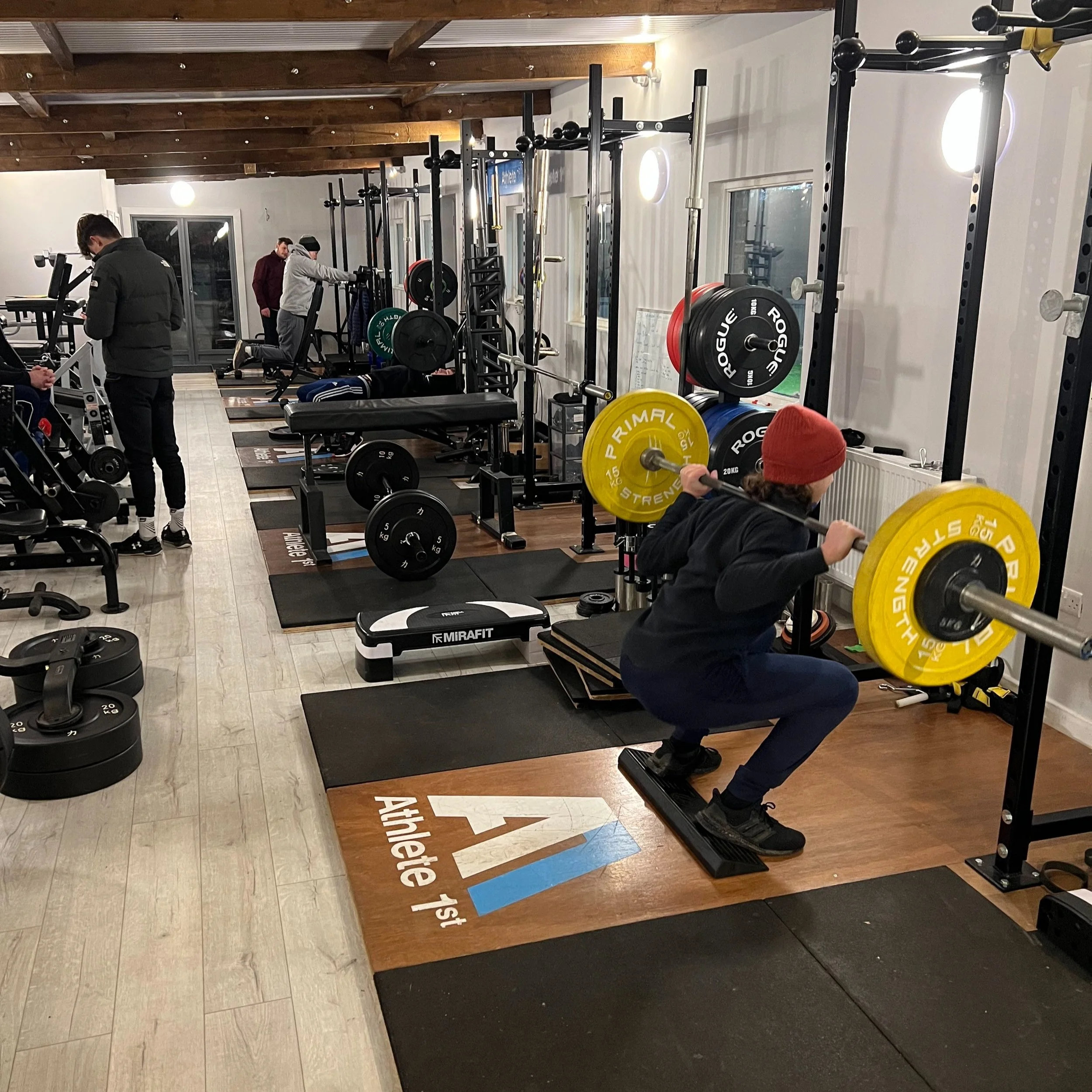 A gym with various weightlifting equipment and people exercising, including a person performing a squat with a barbell loaded with yellow and black weights.