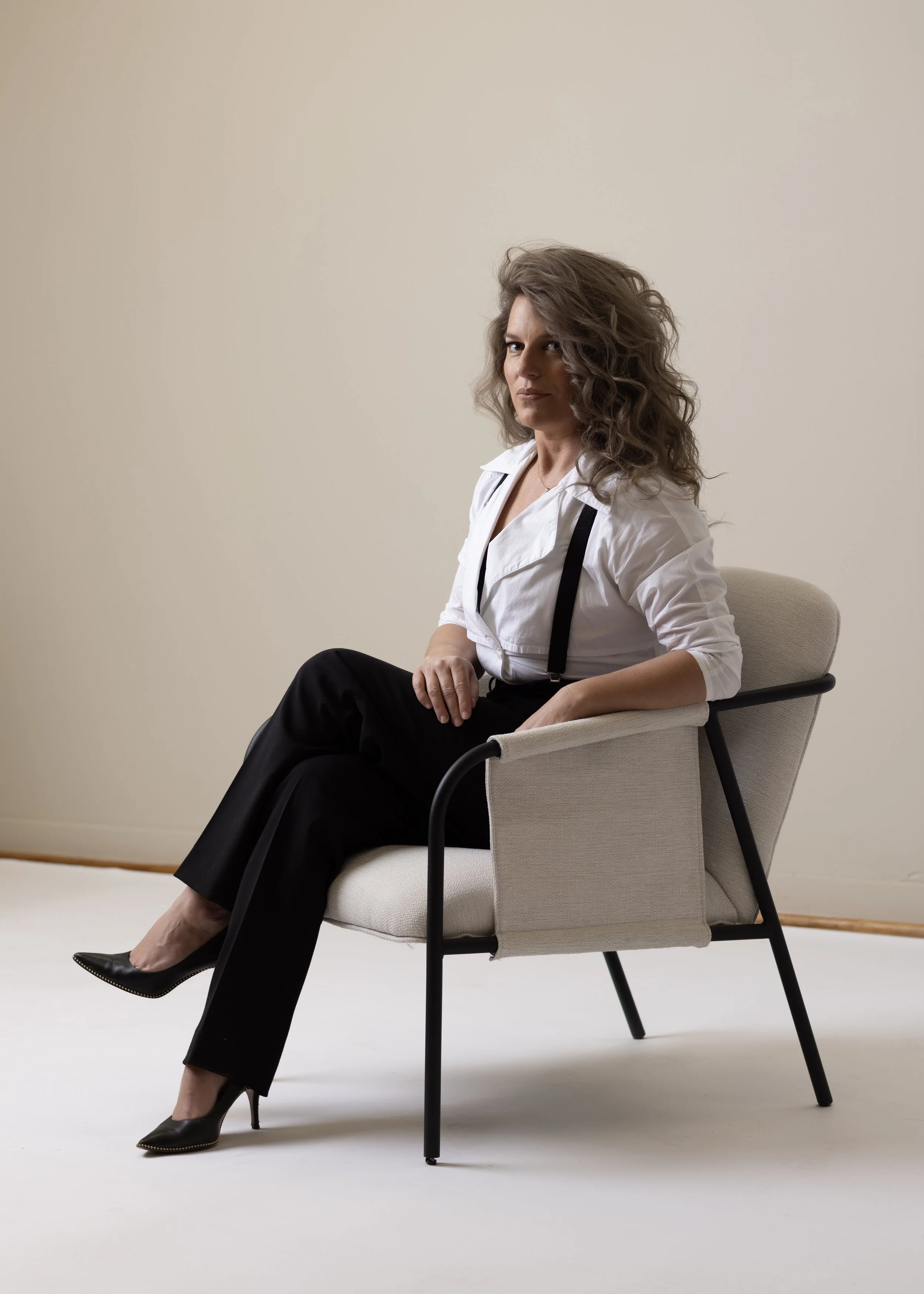 A person with curly hair sitting in a modern chair, wearing a white shirt with suspenders, black pants, and high heels. Headshot and personal branding Fayetteville, AR.