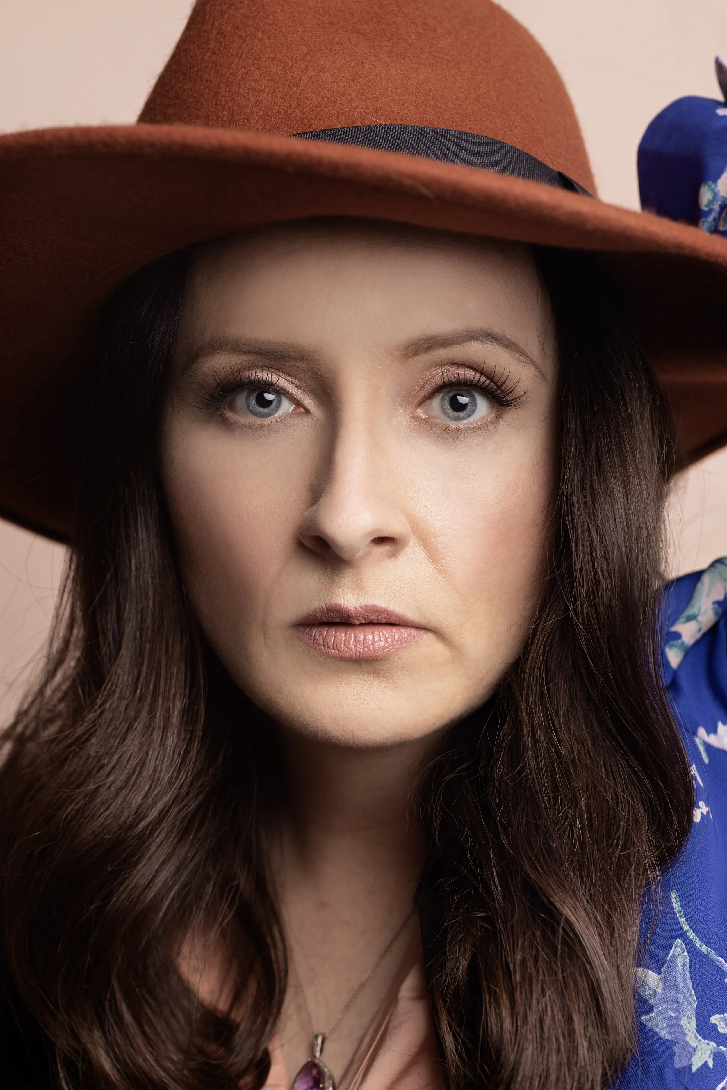 Woman with brown hat and long hair, wearing a blue floral outfit, looking directly at the camera. Headshot and personal branding Fayetteville, AR.