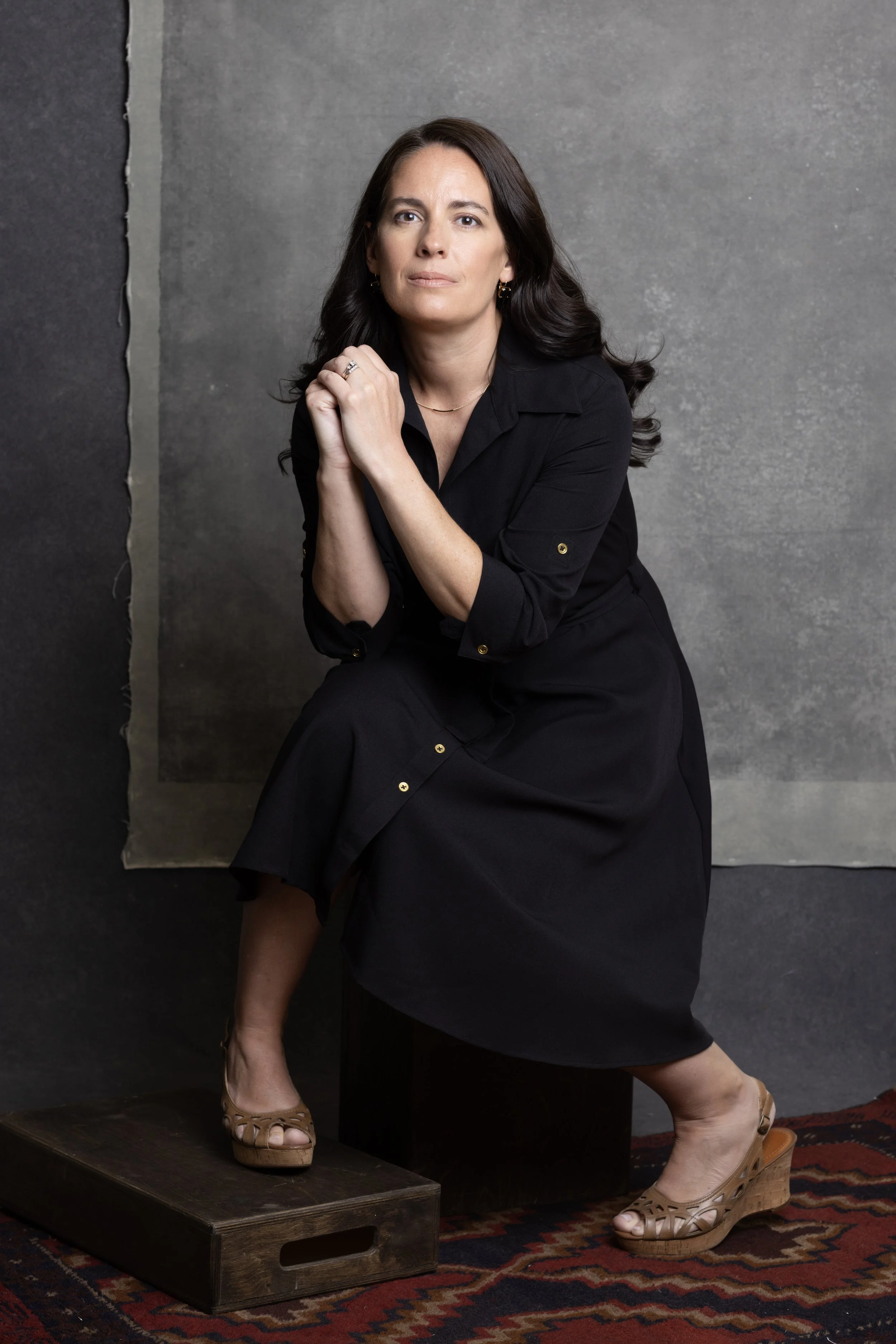 Woman in black dress sitting on wooden block with a thoughtful expression. Headshot and personal branding Fayetteville, AR. Family Portratis.
