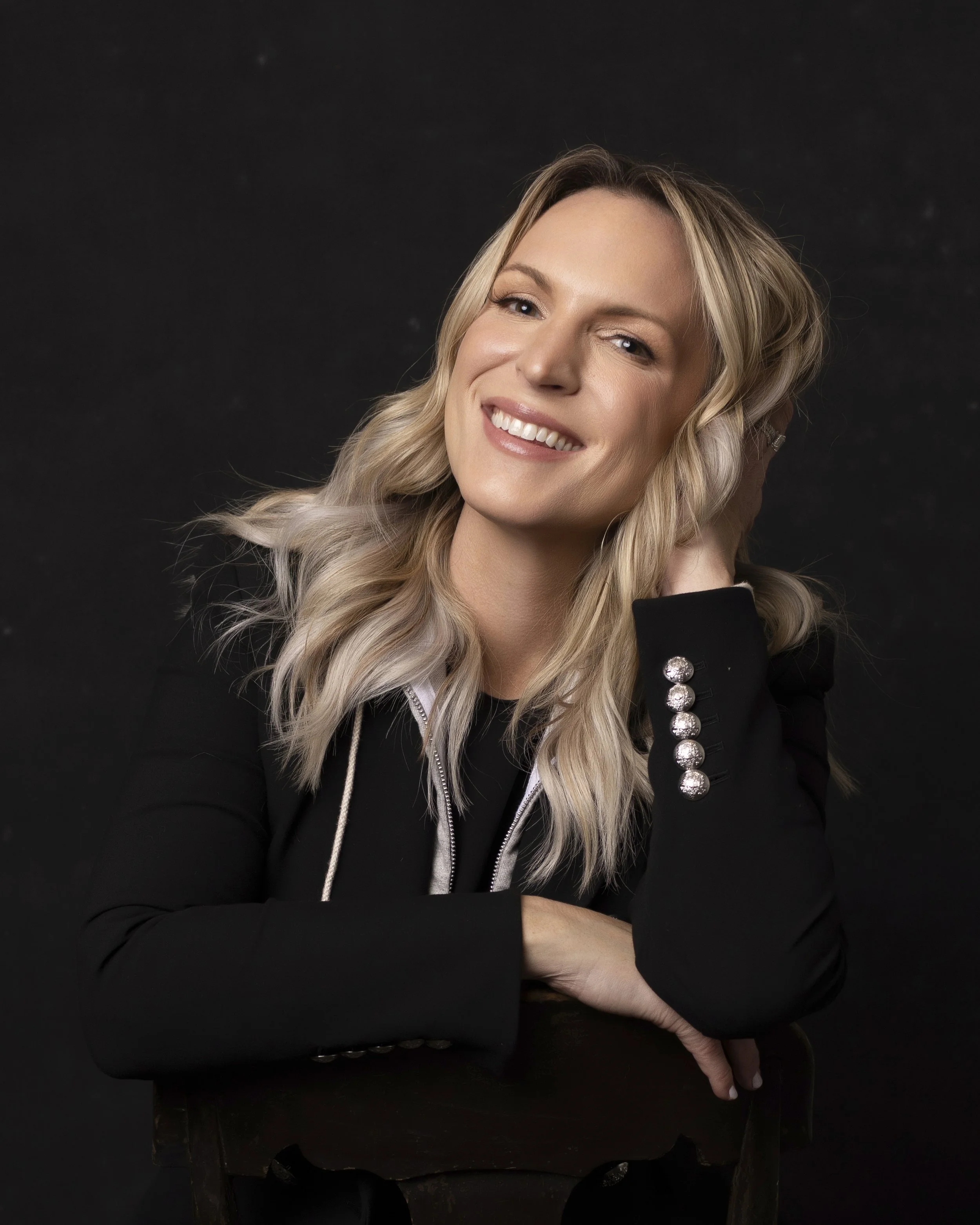 Portrait of a smiling woman with blonde hair, wearing a black jacket with silver buttons, posing against a dark background. Bentonville AR