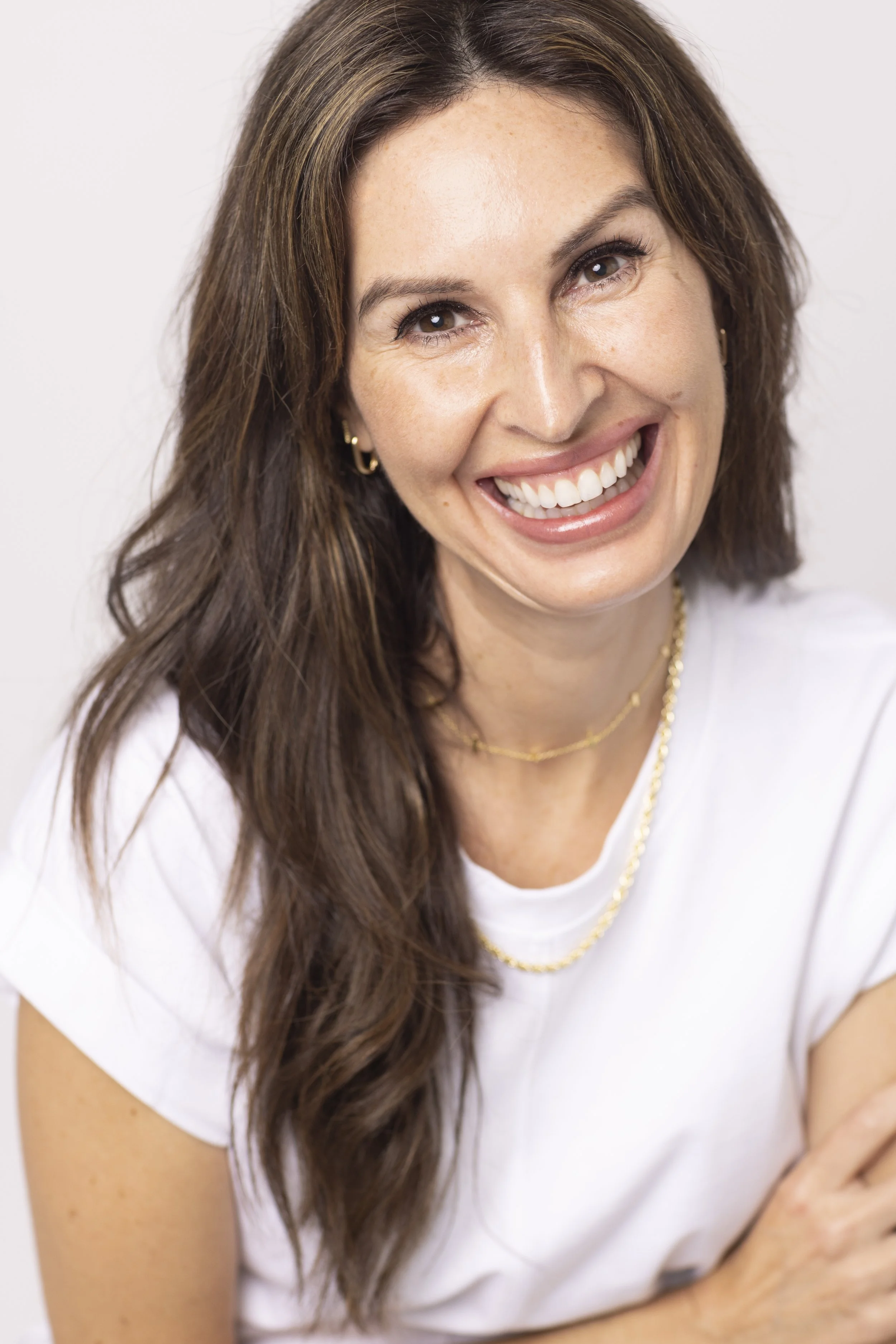 Smiling woman with long dark hair wearing a white shirt and gold necklaces. Headshot and personal branding 