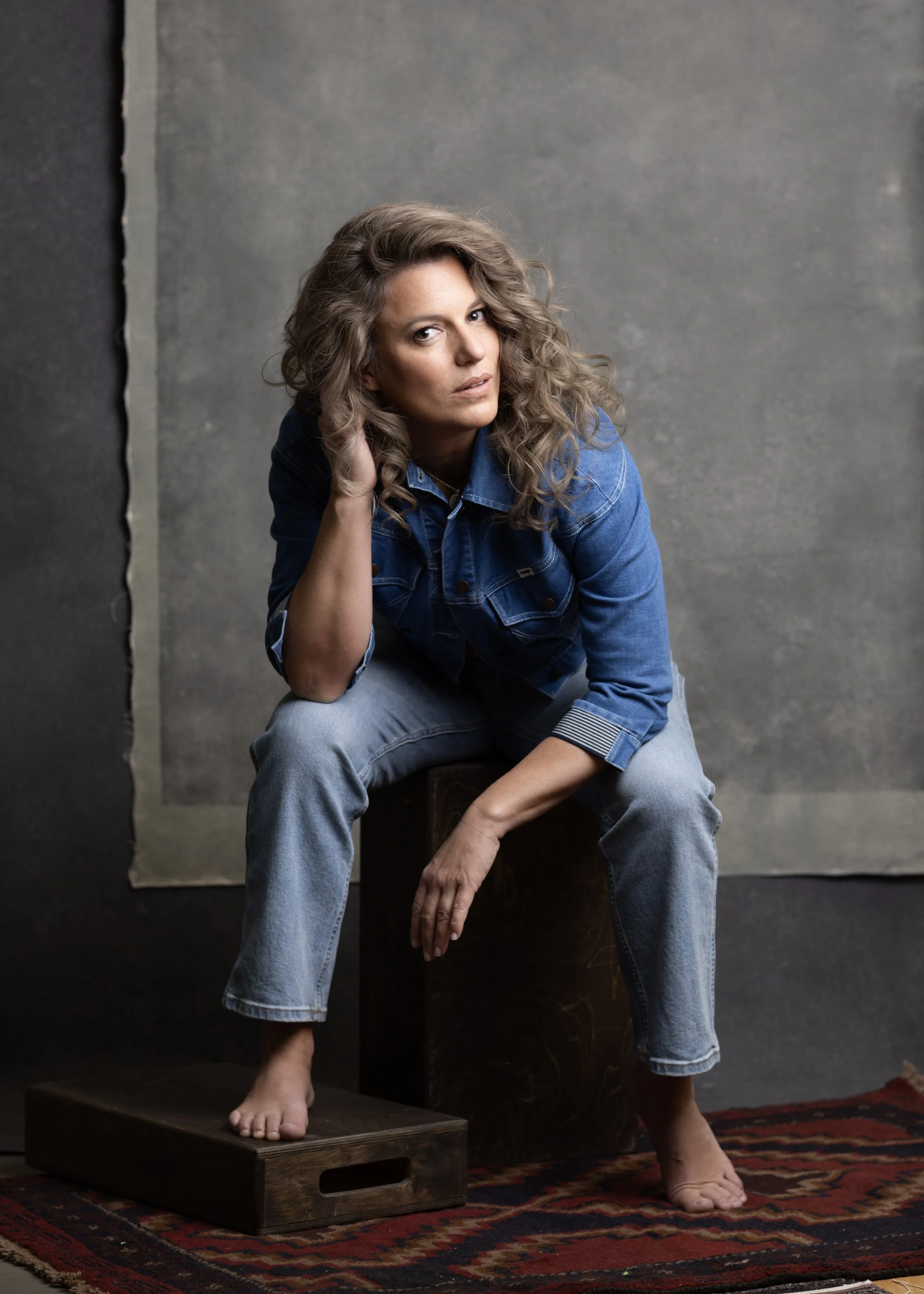 Woman with curly hair wearing a denim jacket and jeans, sitting barefoot on a wooden box with a patterned rug and gray background. Headshot and personal branding Fayetteville, AR.