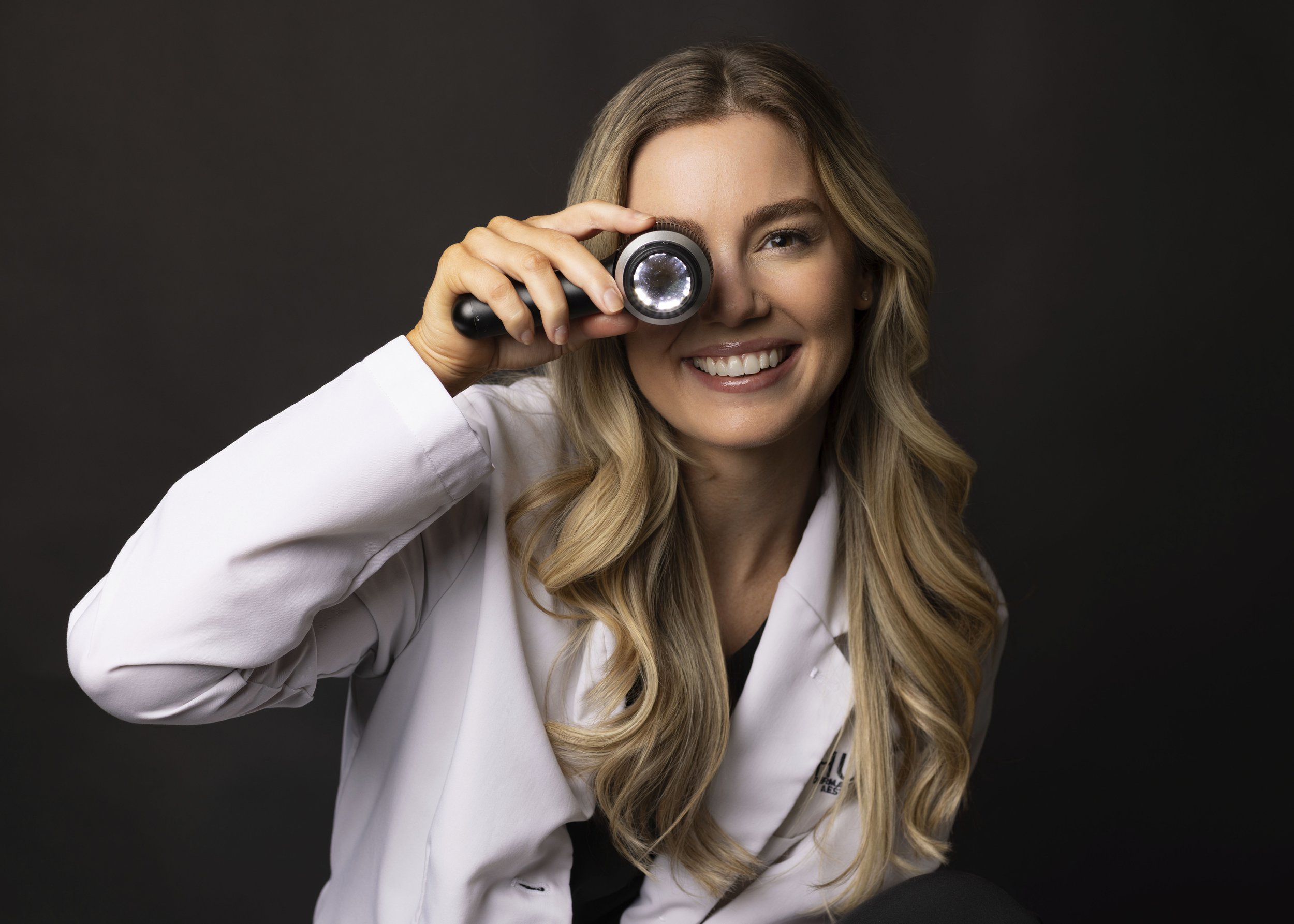 A woman with long blonde hair smiling while holding a flashlight up to her eye, wearing a white lab coat, against a dark background. White Coat Collection Fayetteville Arkansas