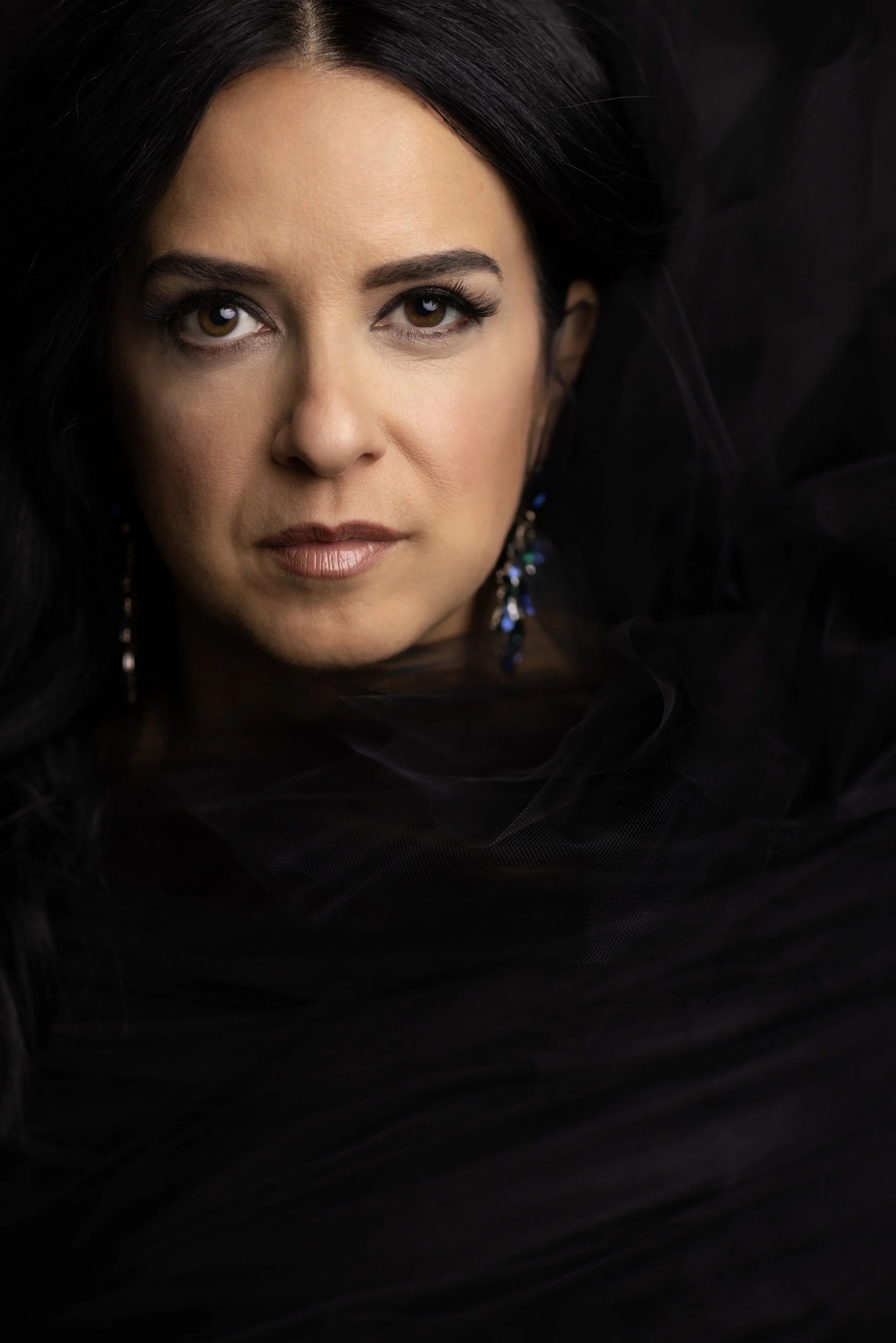 Close-up of a woman with black hair and dark clothing, wearing earrings against a dark background. Headshot and personal branding Fayetteville, AR.