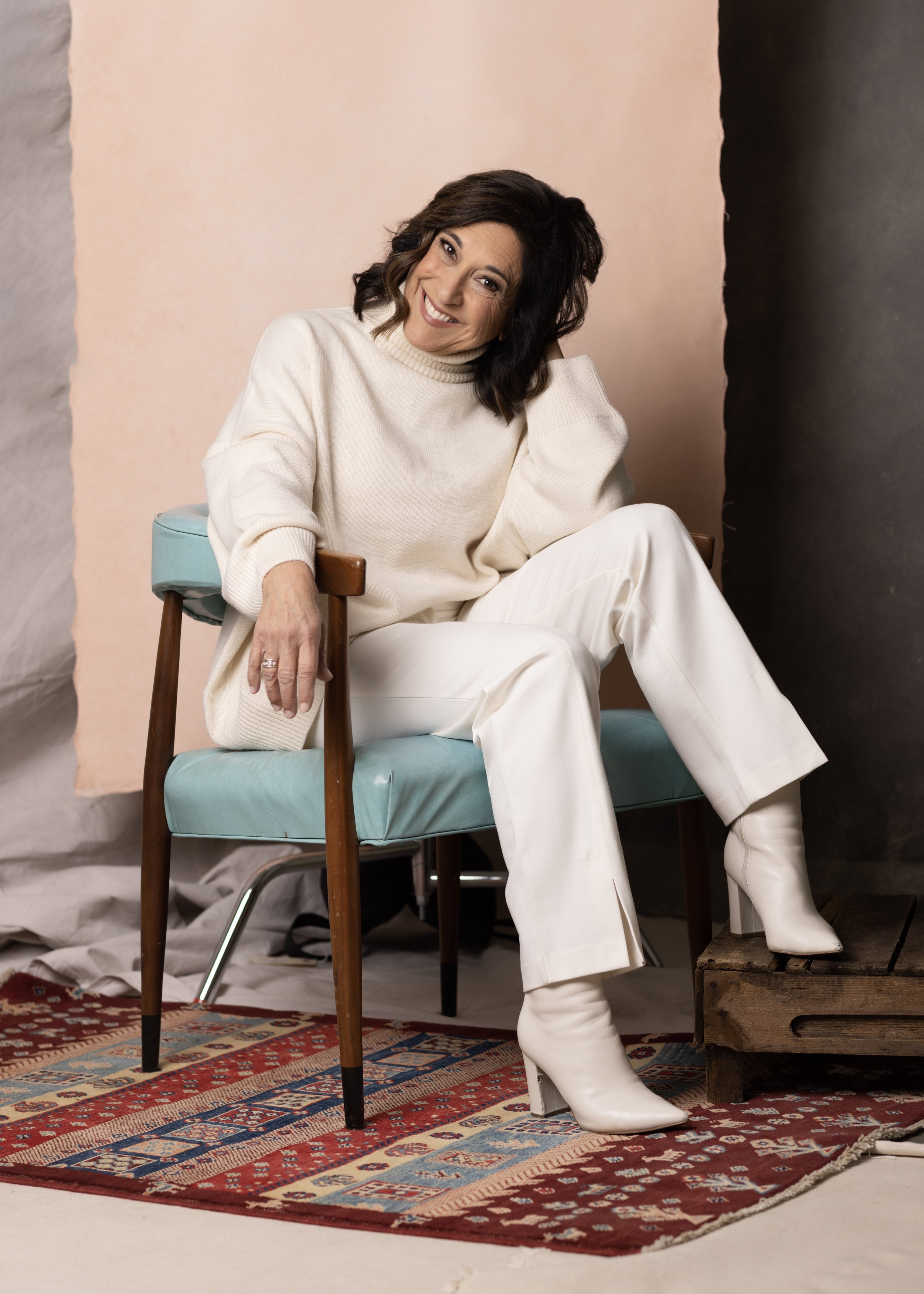 A woman sitting on a turquoise armchair, wearing a white sweater, white pants, and white boots, smiling against a backdrop with a patterned rug on the floor. Headshot and personal branding Fayetteville, AR.