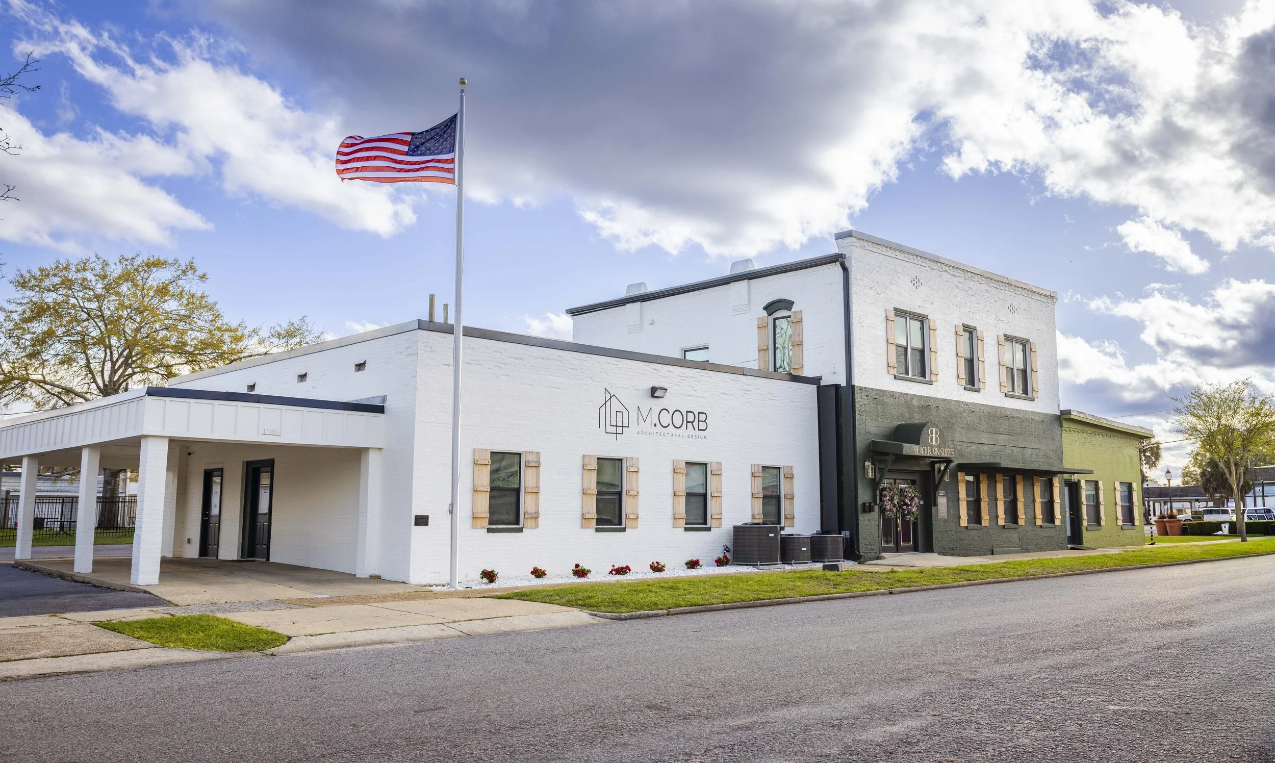 A two-story building with white and dark gray exterior walls, signage for architectural and interior design companies, an American flag on a flagpole, and trees in the background under a partly cloudy sky.