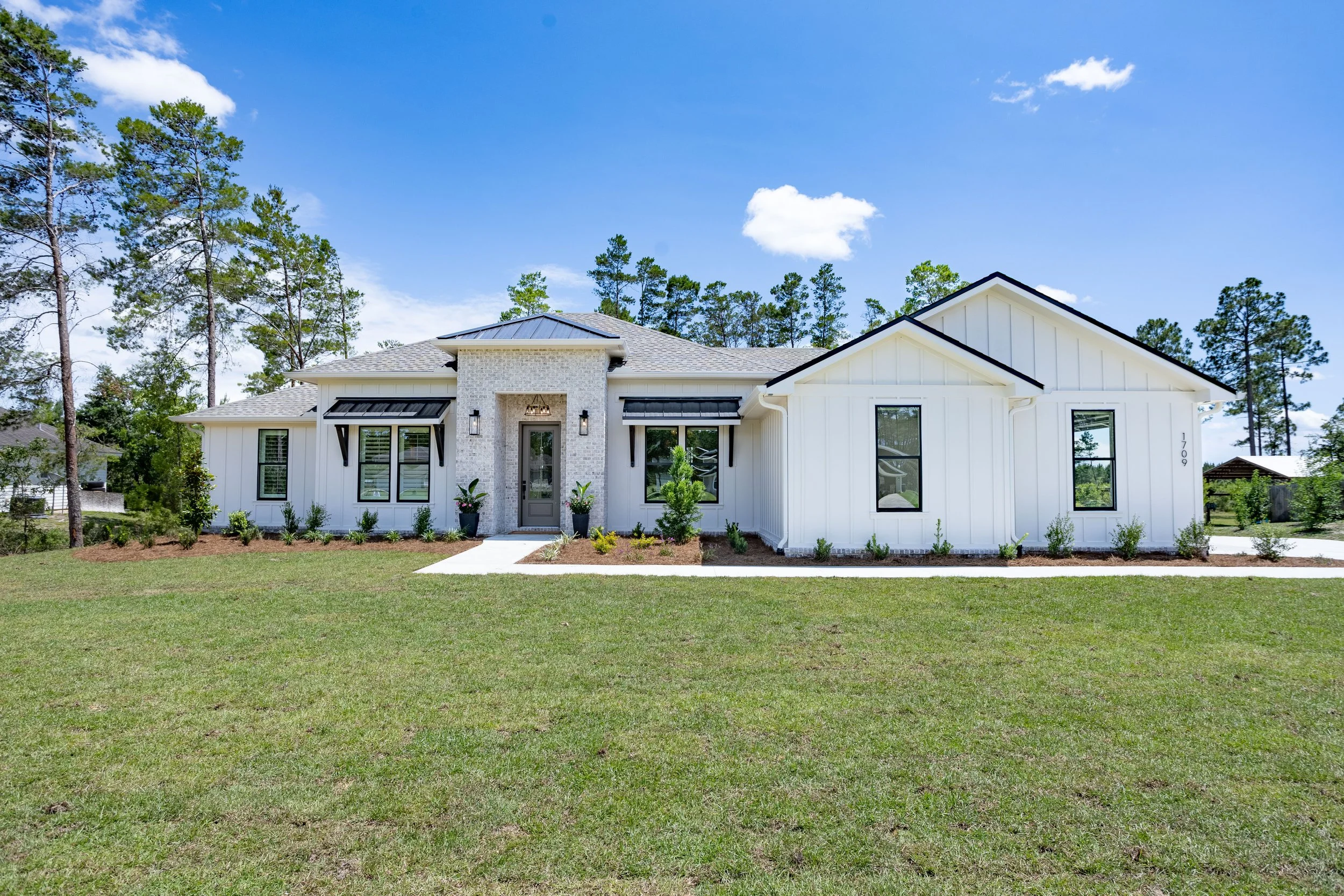 White modern house with black metal awnings, landscaped front yard, and a concrete pathway leading to the front door, under a partly cloudy blue sky.