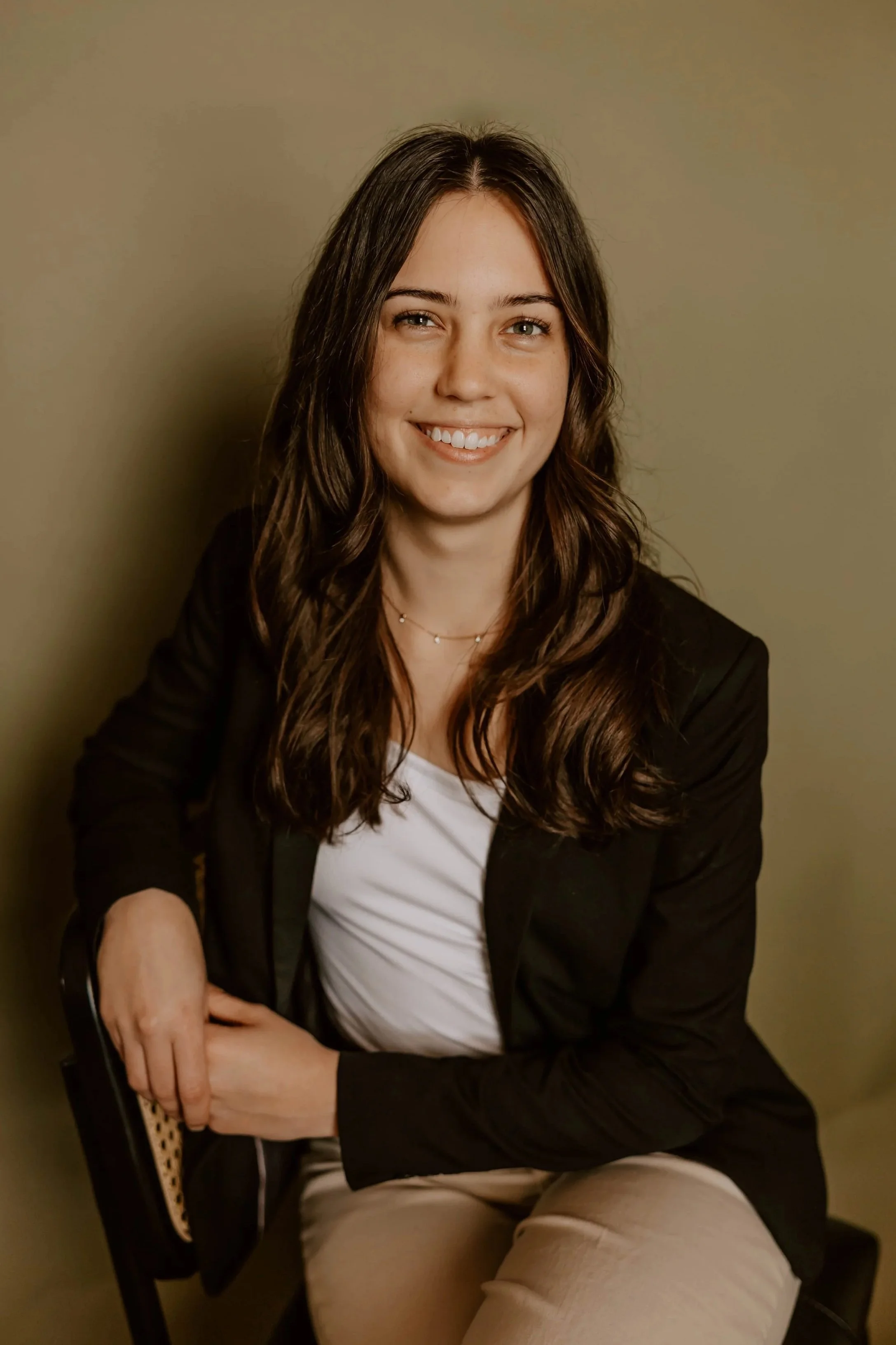A smiling woman with long brown hair, wearing a black jacket over a white top, beige pants, sitting with crossed arms against a plain beige wall.