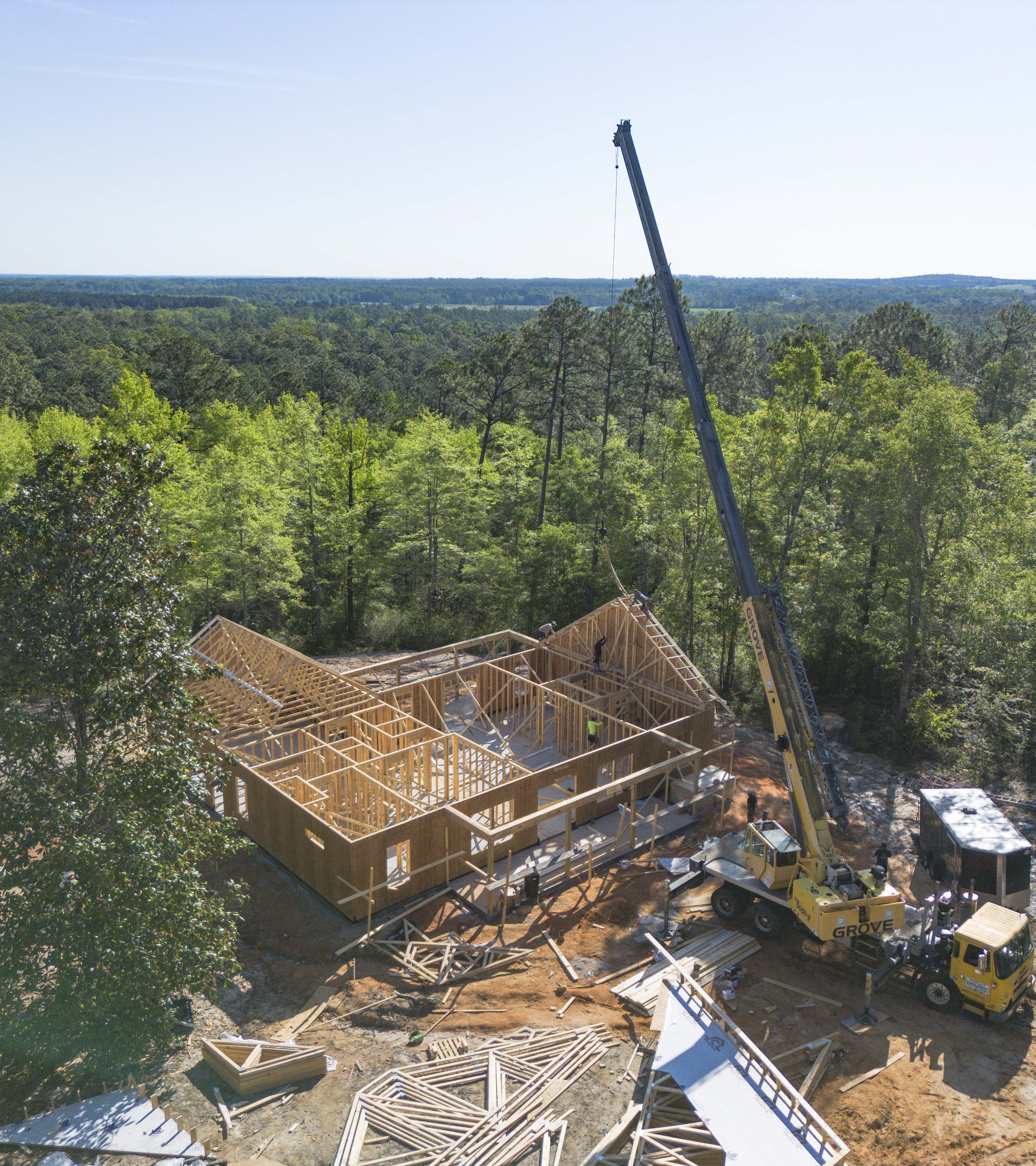 Construction site with a wooden framework for a building, a large crane lifting materials, and construction workers working on the structure, surrounded by trees and forested area.