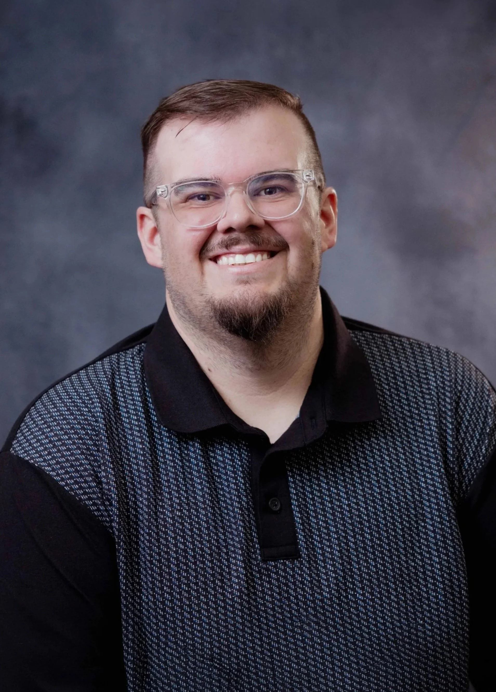 A smiling man with short brown hair, glasses, and a beard, wearing a black collared shirt with a patterned vest, against a dark, cloudy background.