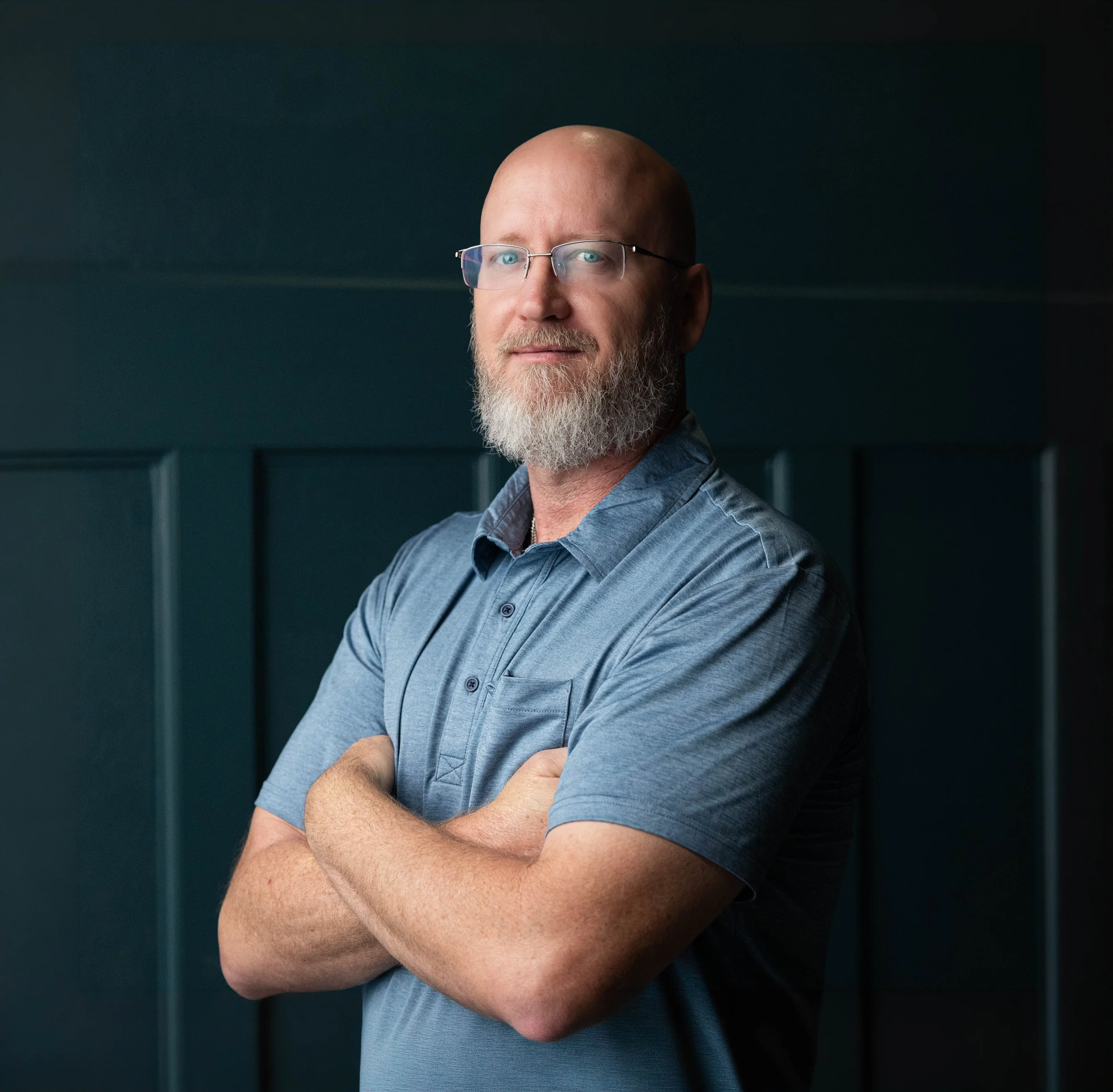 A middle-aged man with a beard and glasses, wearing a blue shirt, crossing his arms and looking confidently at the camera, standing against a dark background.