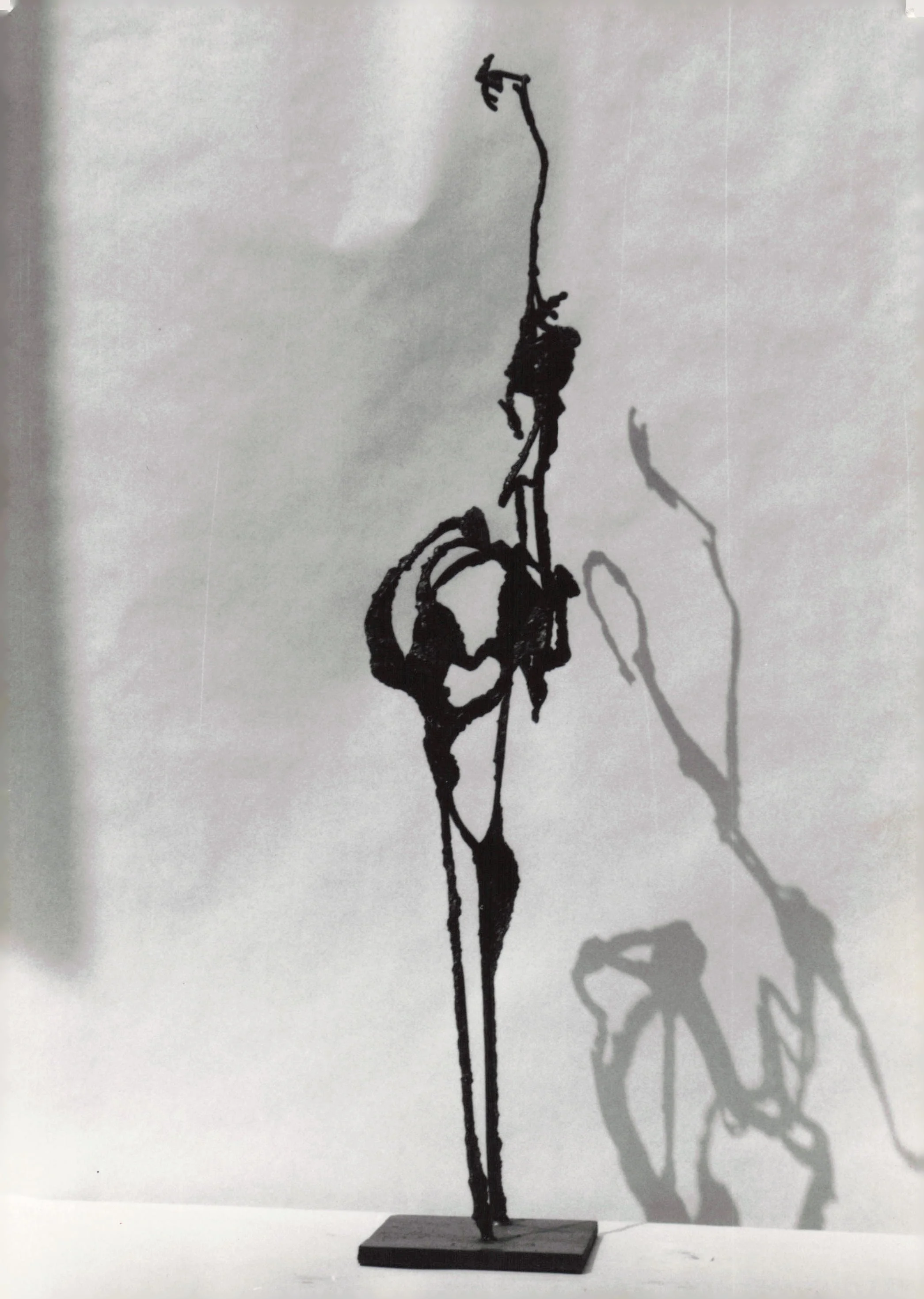 Sculpture of a tall, thin, abstract figure with elongated limbs, standing on a wooden base, casting a shadow on the wall behind.