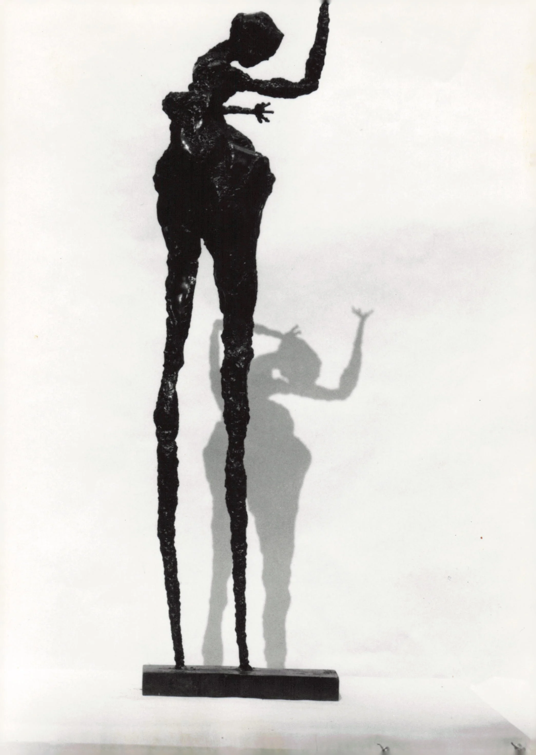 Metal sculpture of a tall, thin figure with elongated limbs, bending forward while holding a long object, with a small object or animal at its feet, cast shadows on a white background.