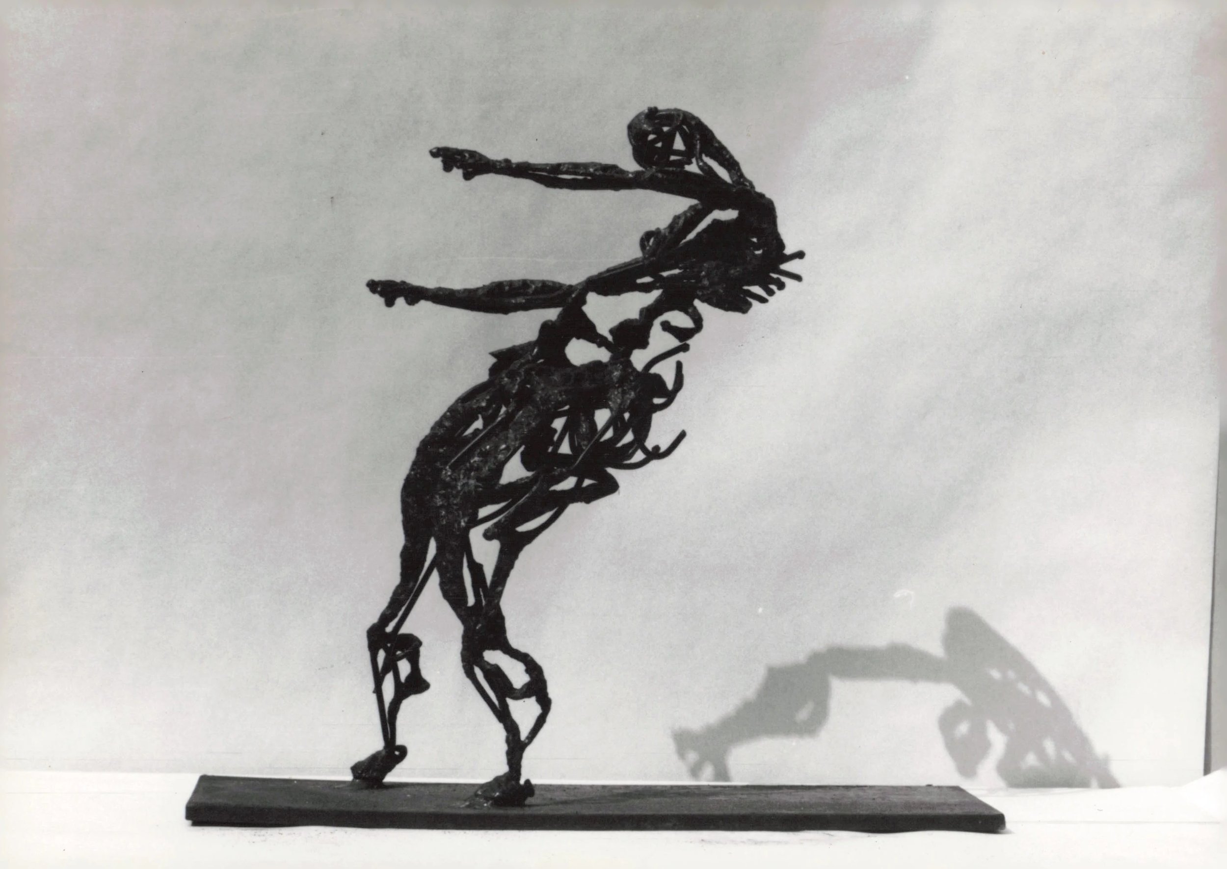 Black wire sculpture of a girl pointing, mounted on a black base, casting a shadow on a white background.