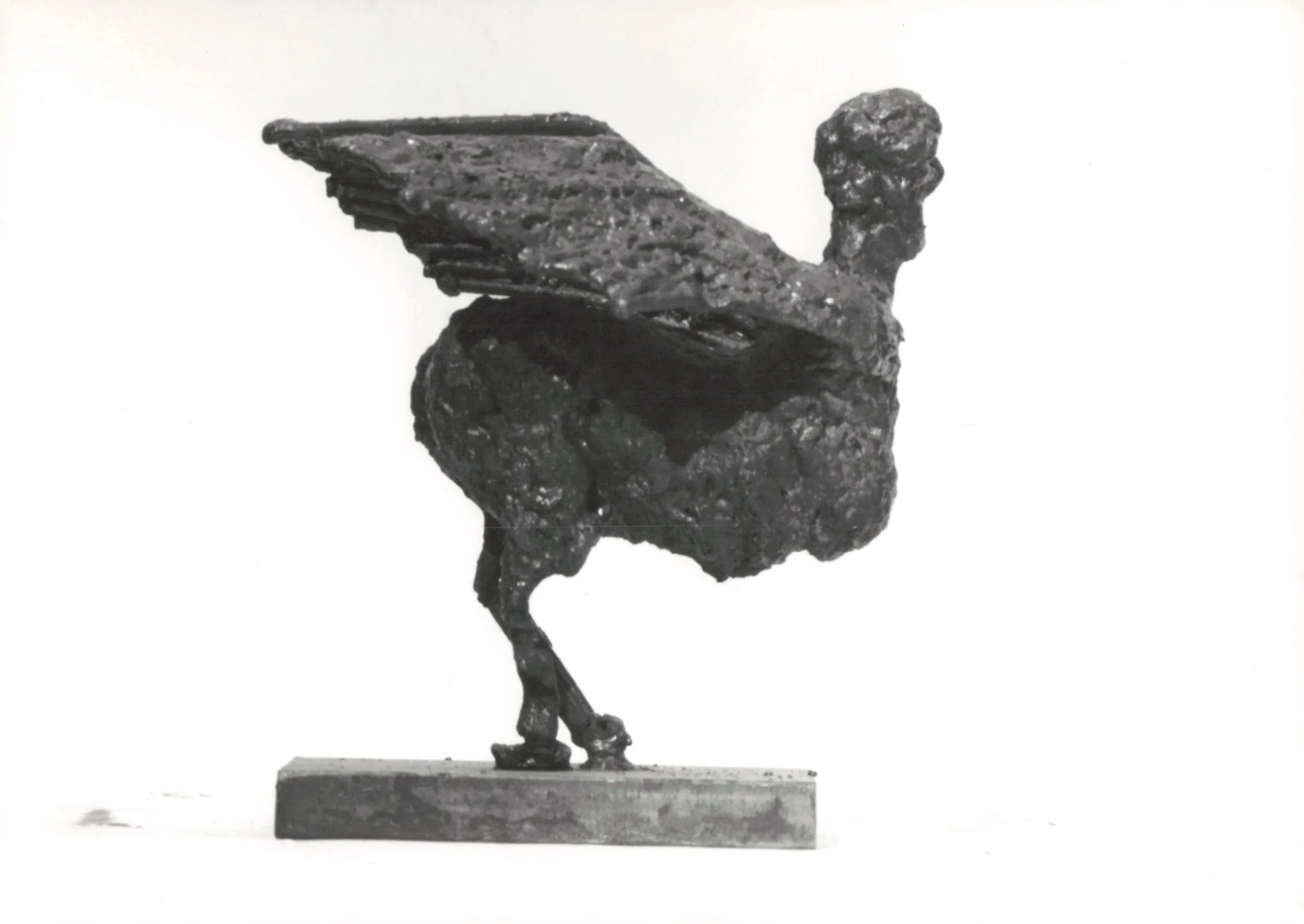 A black and white sculpture of a bird standing on one leg, with its wings slightly raised and head turned to the side, mounted on a rectangular base.
