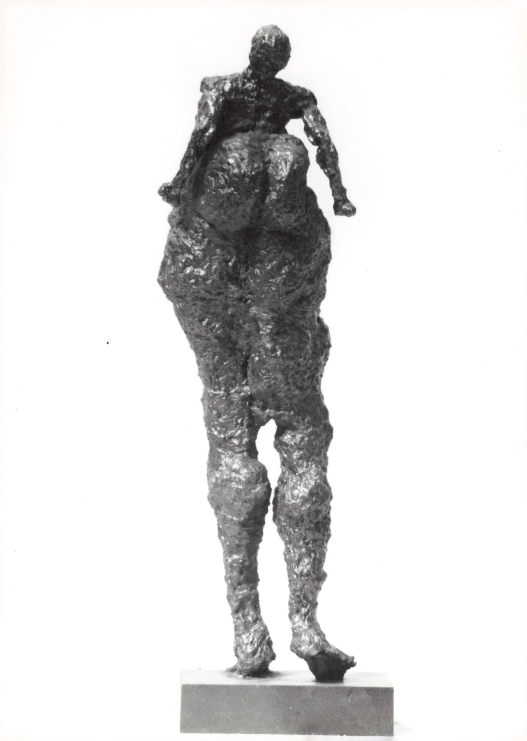 A sculpture of a person standing on a rectangular base, with their back turned and arms relaxed at their sides.