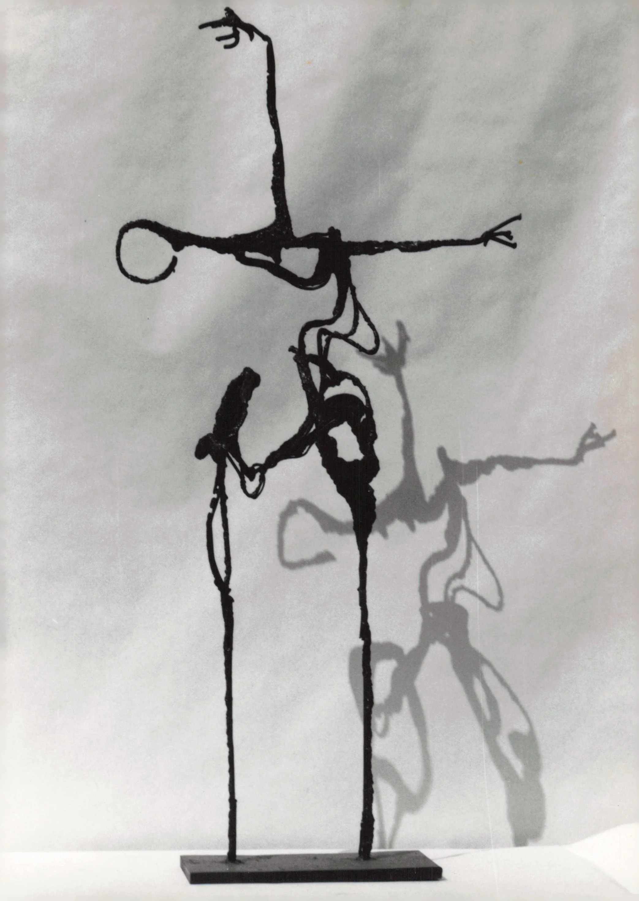 Abstract black wire sculpture of two human figures on a rectangular base, casting shadows on a white background.