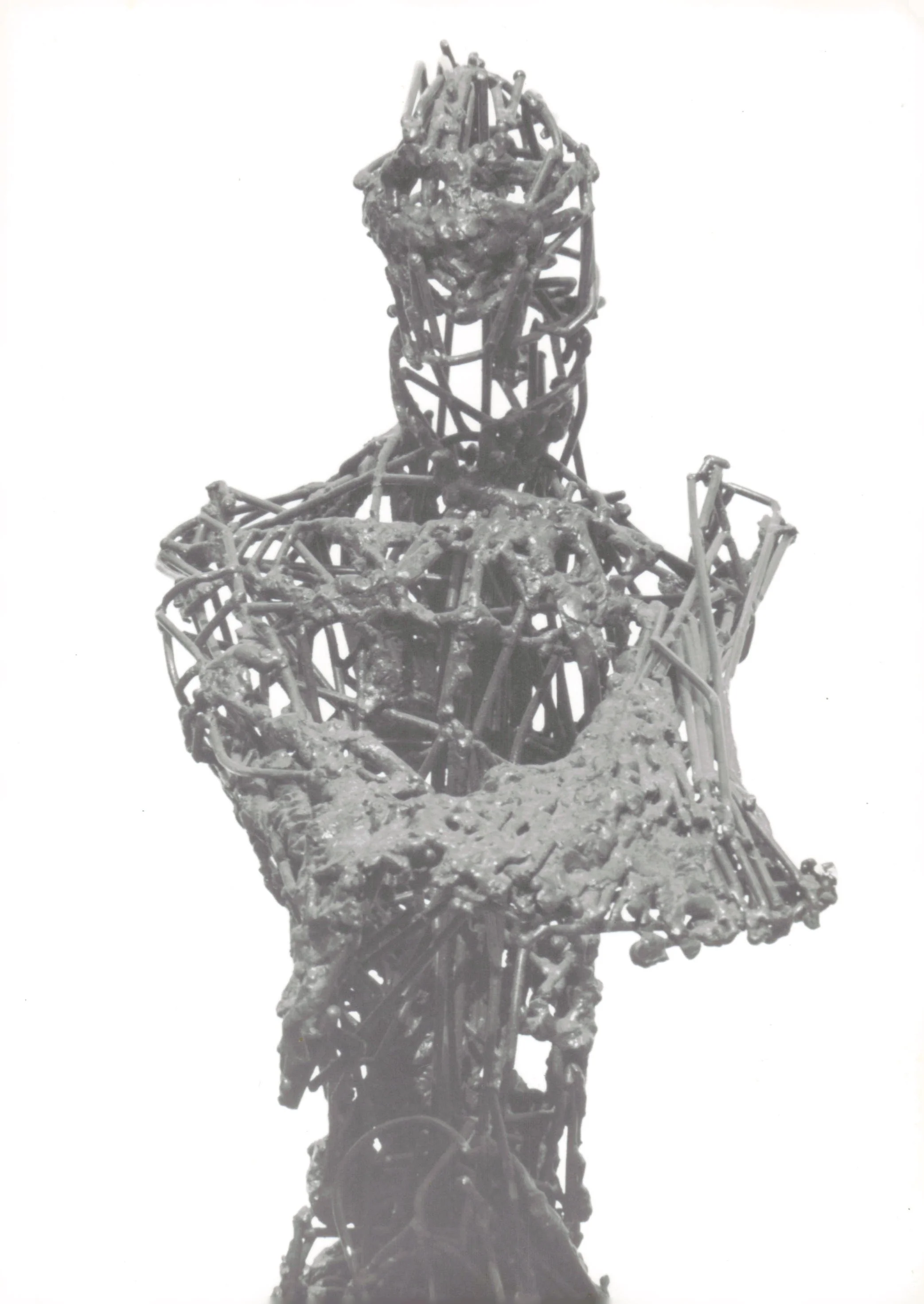 Sculpture of a human figure made from intertwined metal rods, in a seated pose with a flat, textured ground.