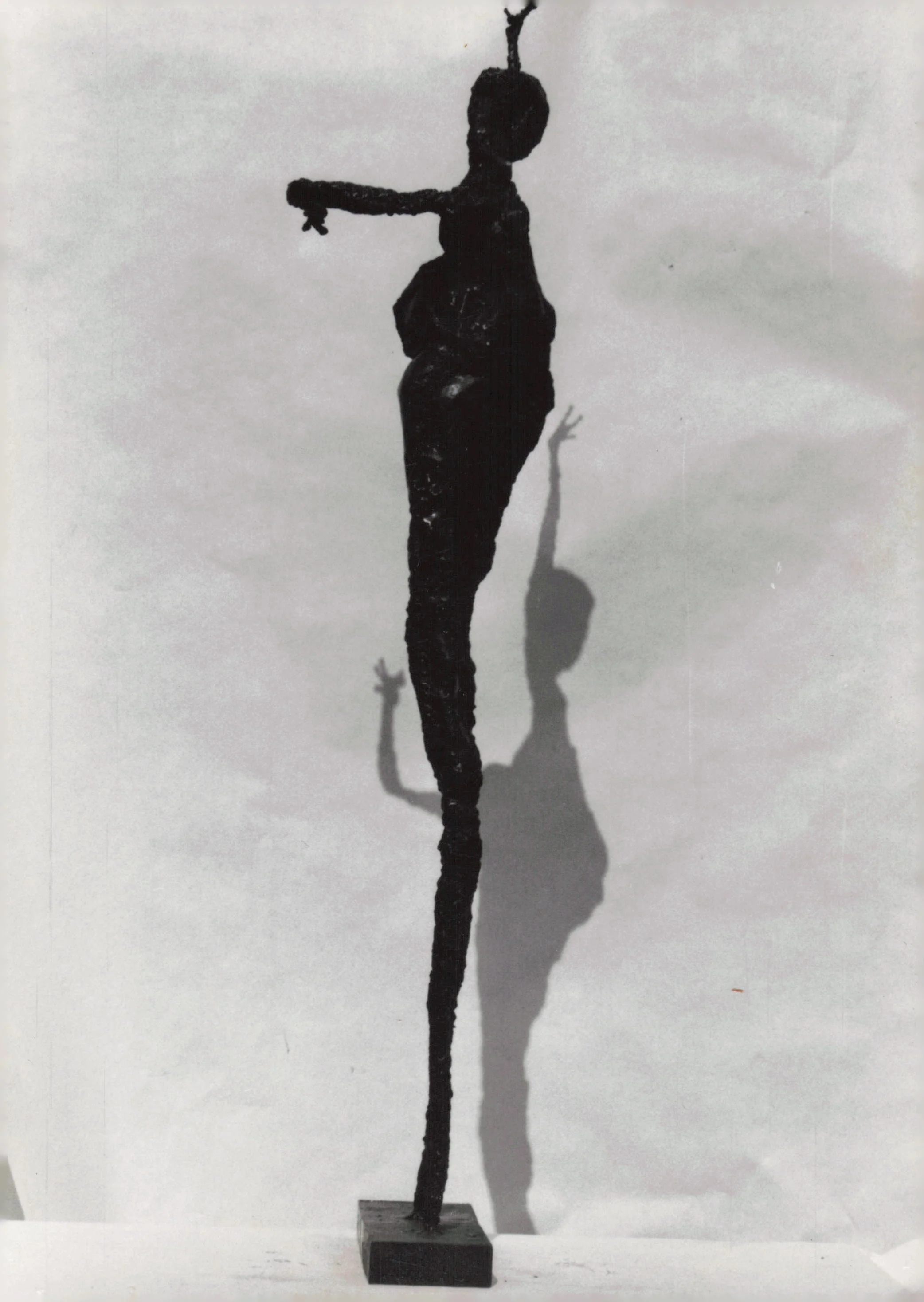 A black metal sculpture of a woman with one arm extended, standing on a rectangular base, casting a shadow on a plain wall.