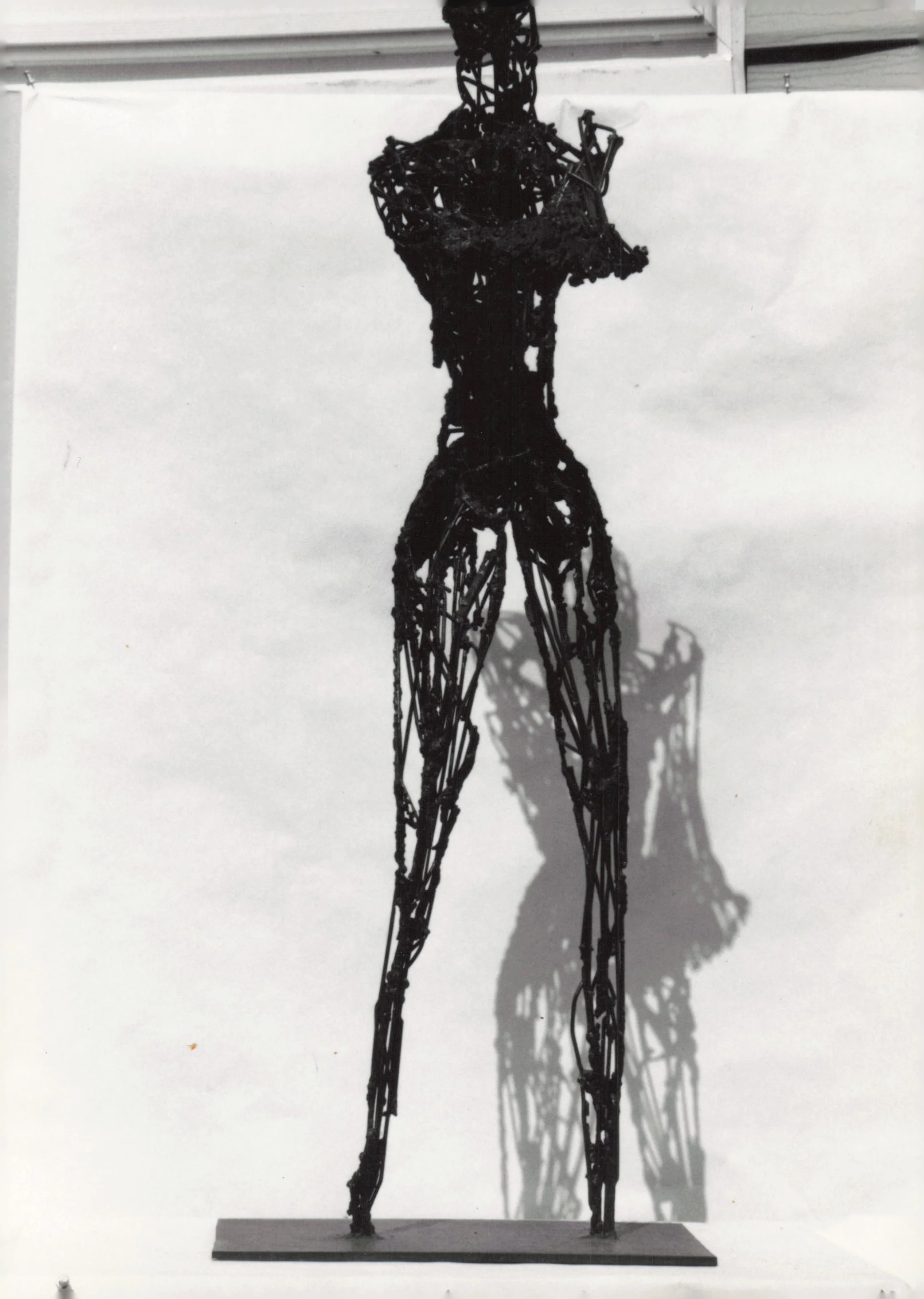 A black metal sculpture of a human figure standing on a rectangular base casts a shadow on the white background.