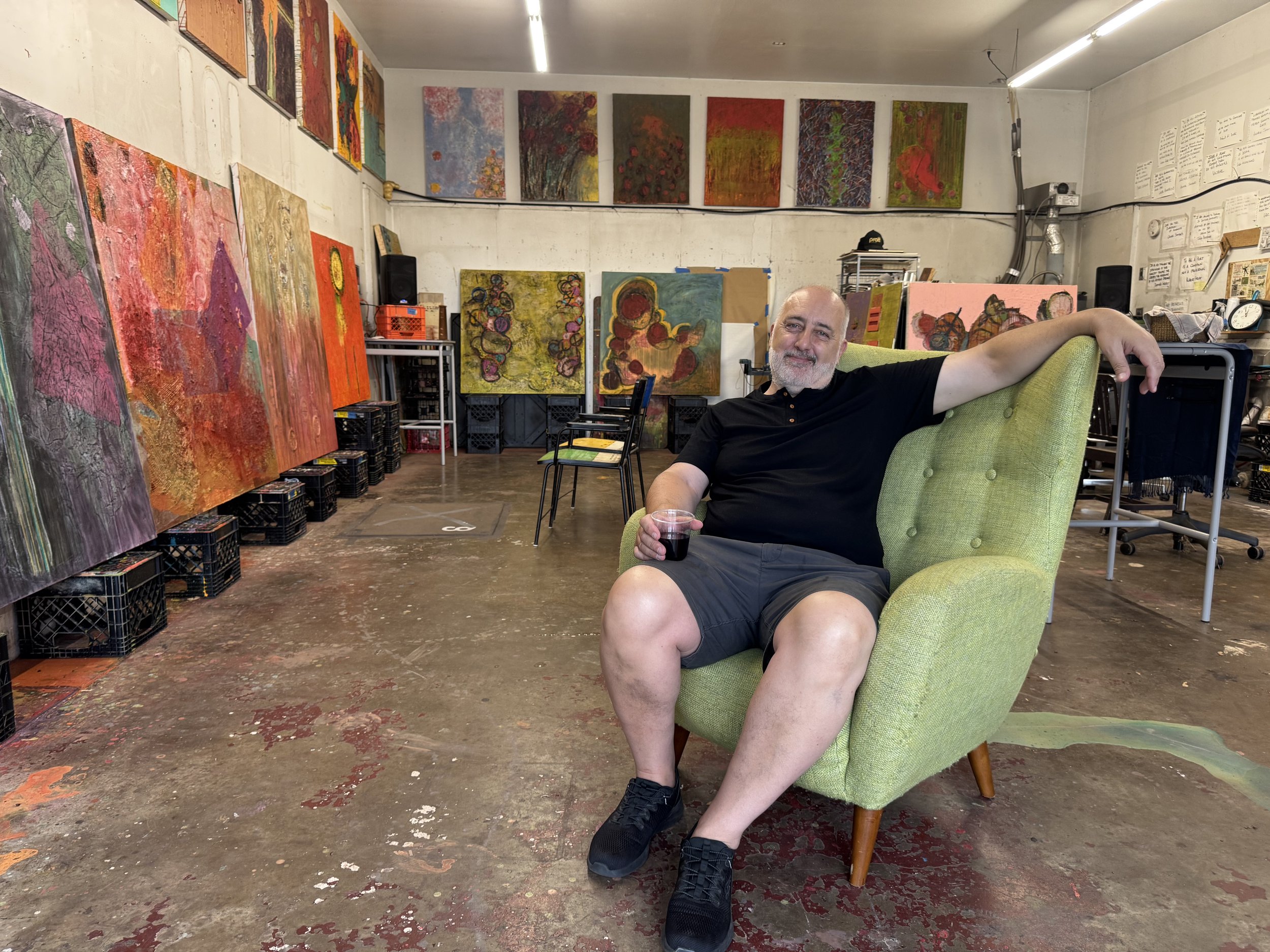 Anthony Scarzafava seated in a green armchair in his art studio, surrounded by his abstract paintings