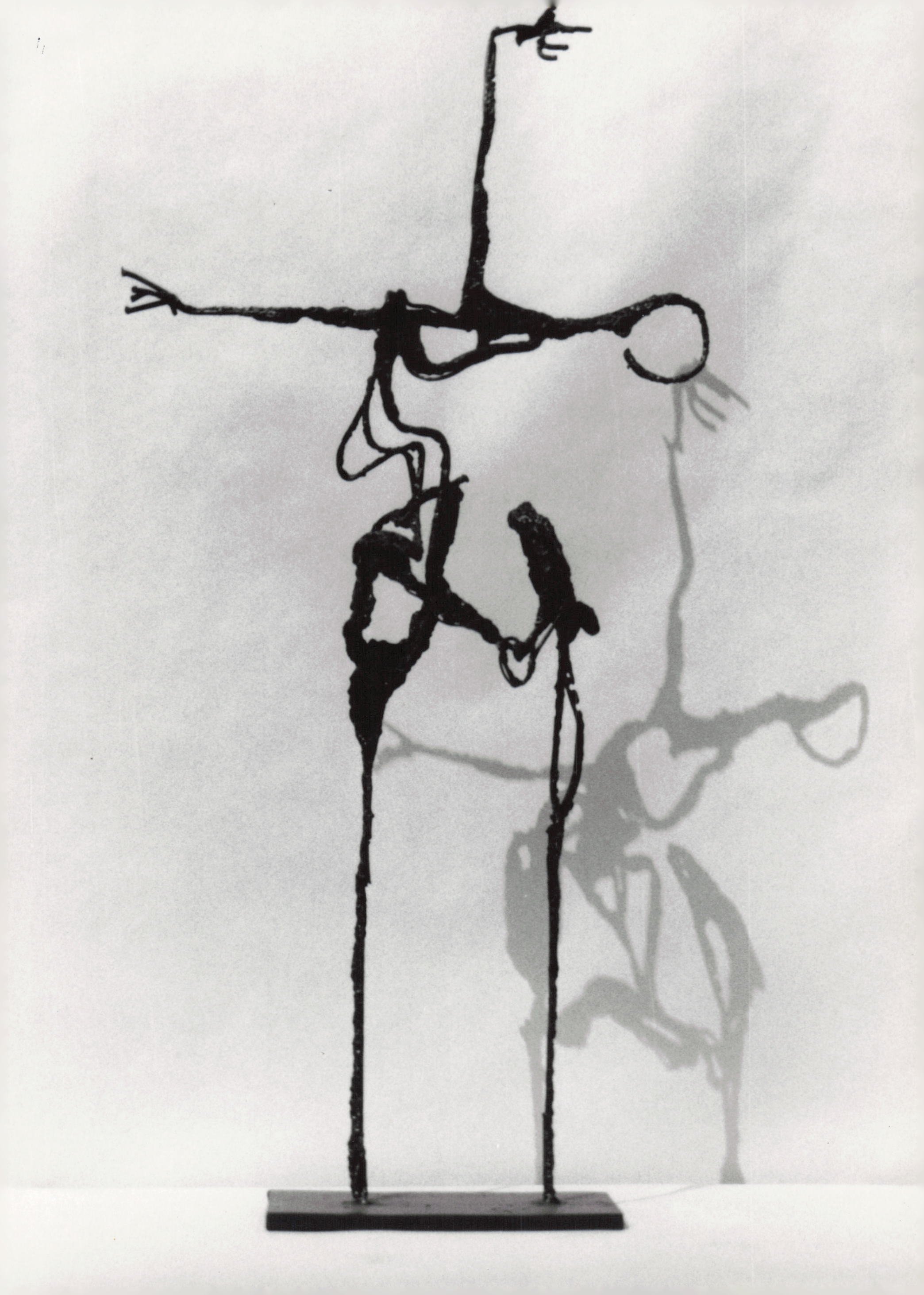 A modern sculpture of a humanoid figure made of metal wires, with elongated limbs and a circular head, standing on a flat base.