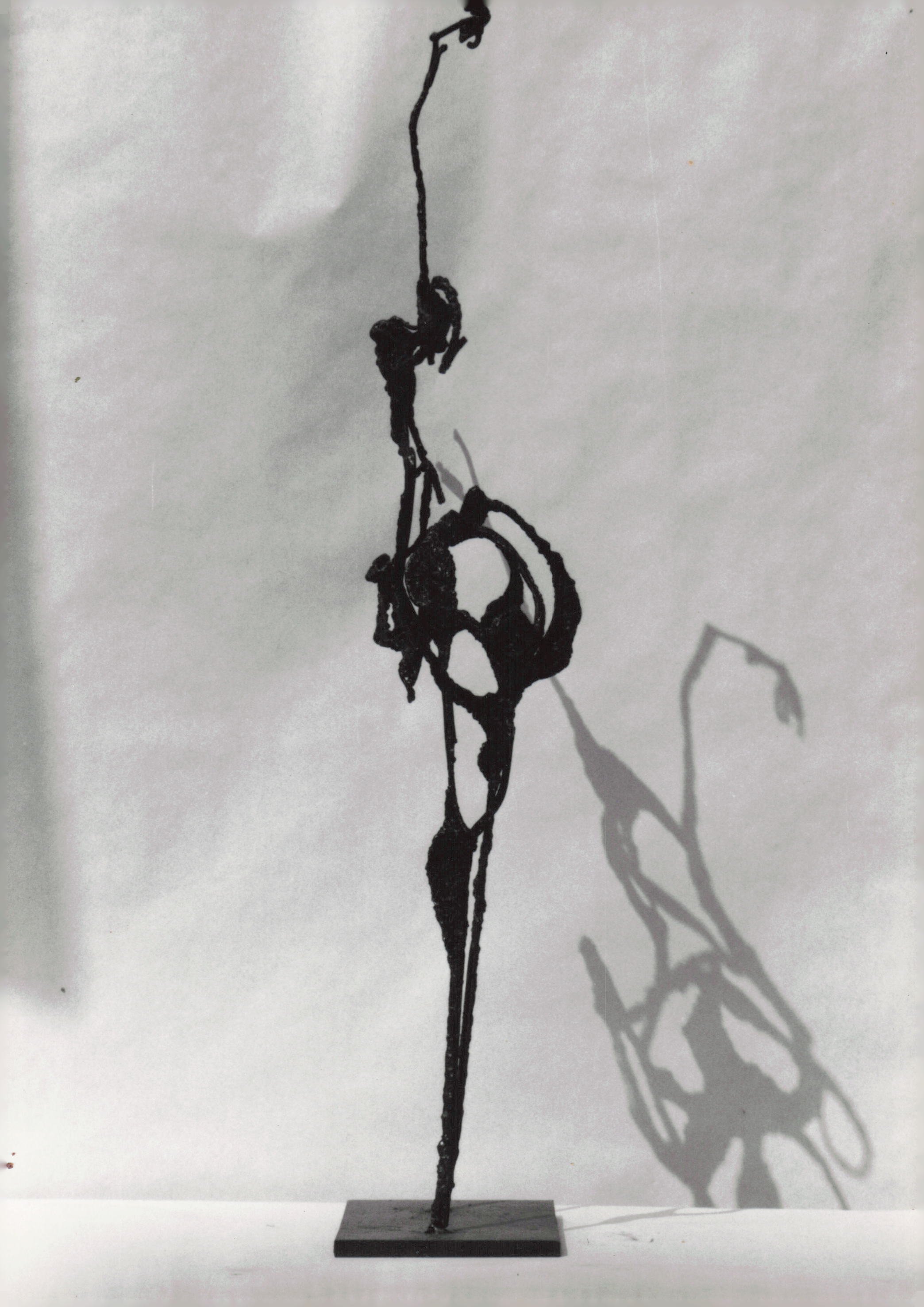 A black wire sculpture of a person holding a pole, mounted on a rectangular base, with its shadow cast on a plain background.