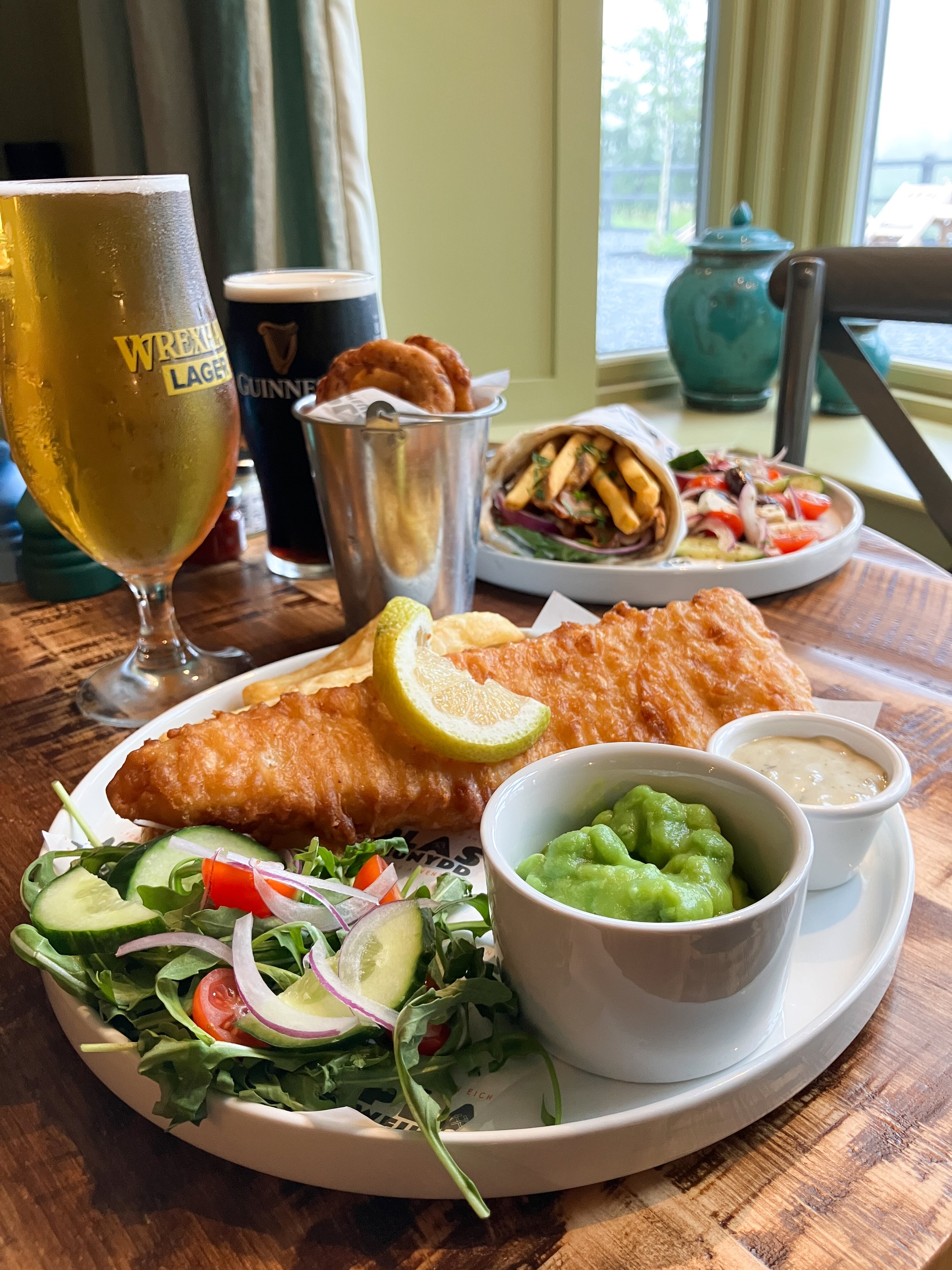 A meal at Plas Weunydd Hotel Restaurant, plate with fried fish, lemon wedges, a side salad with cucumbers, tomatoes, and onions, with tartar sauce and green peas, onion rings, two glasses of beer, one with Wrexham Lager and the other Guinness.