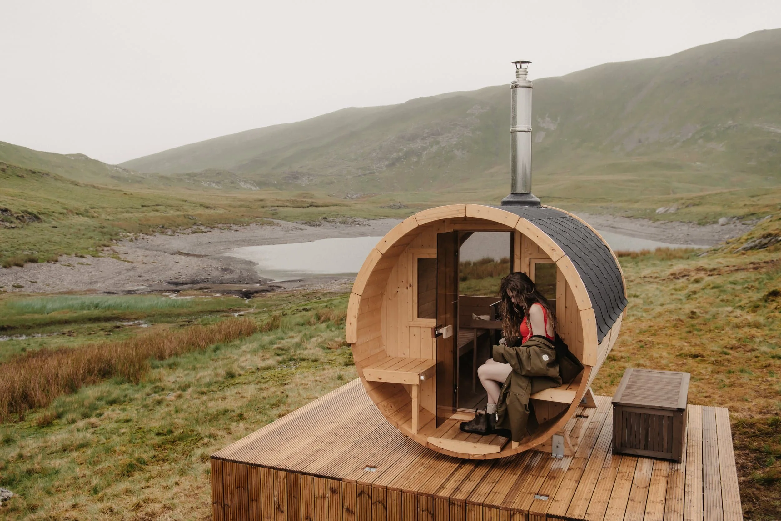 A woman sitting outside of the sauna at Barlwyd. Small, round, wooden sauna in a green, hilly landscape with a lake in the background.