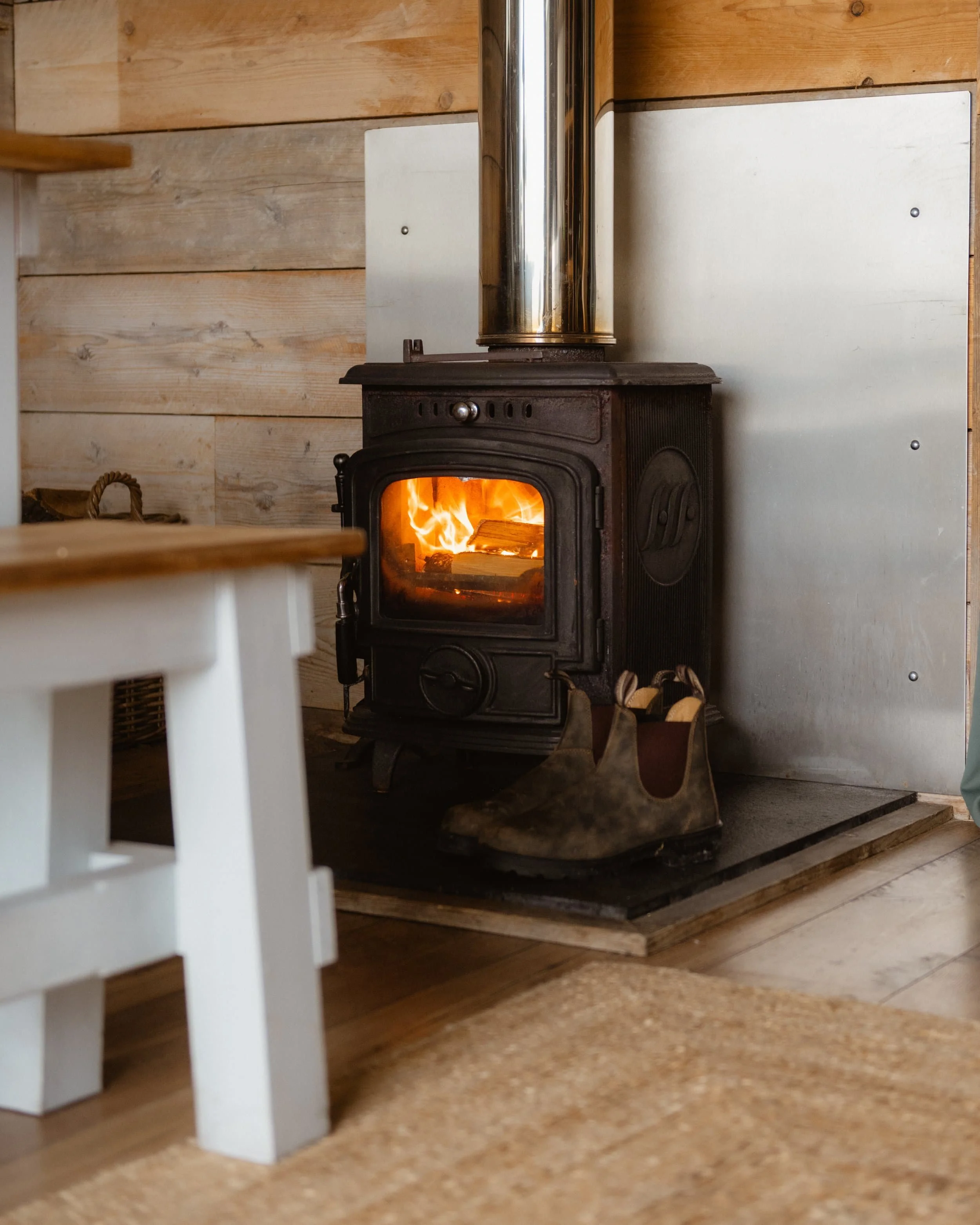 A wood-burning stove with a fire inside, boots in front, in a cosy glamping pod  with rustic decor.